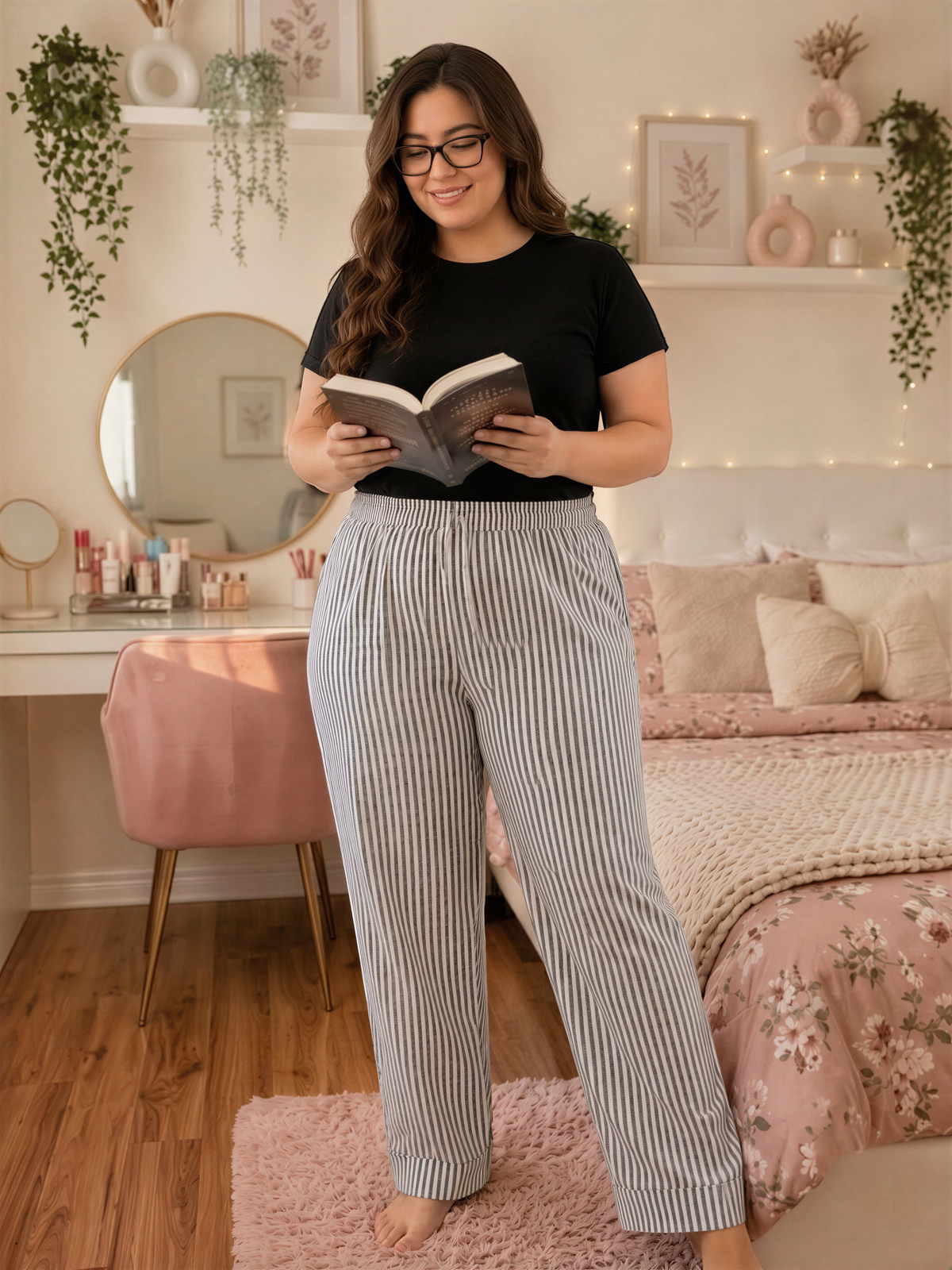 A woman is standing in a bedroom, reading a book while wearing a black top and striped pants.