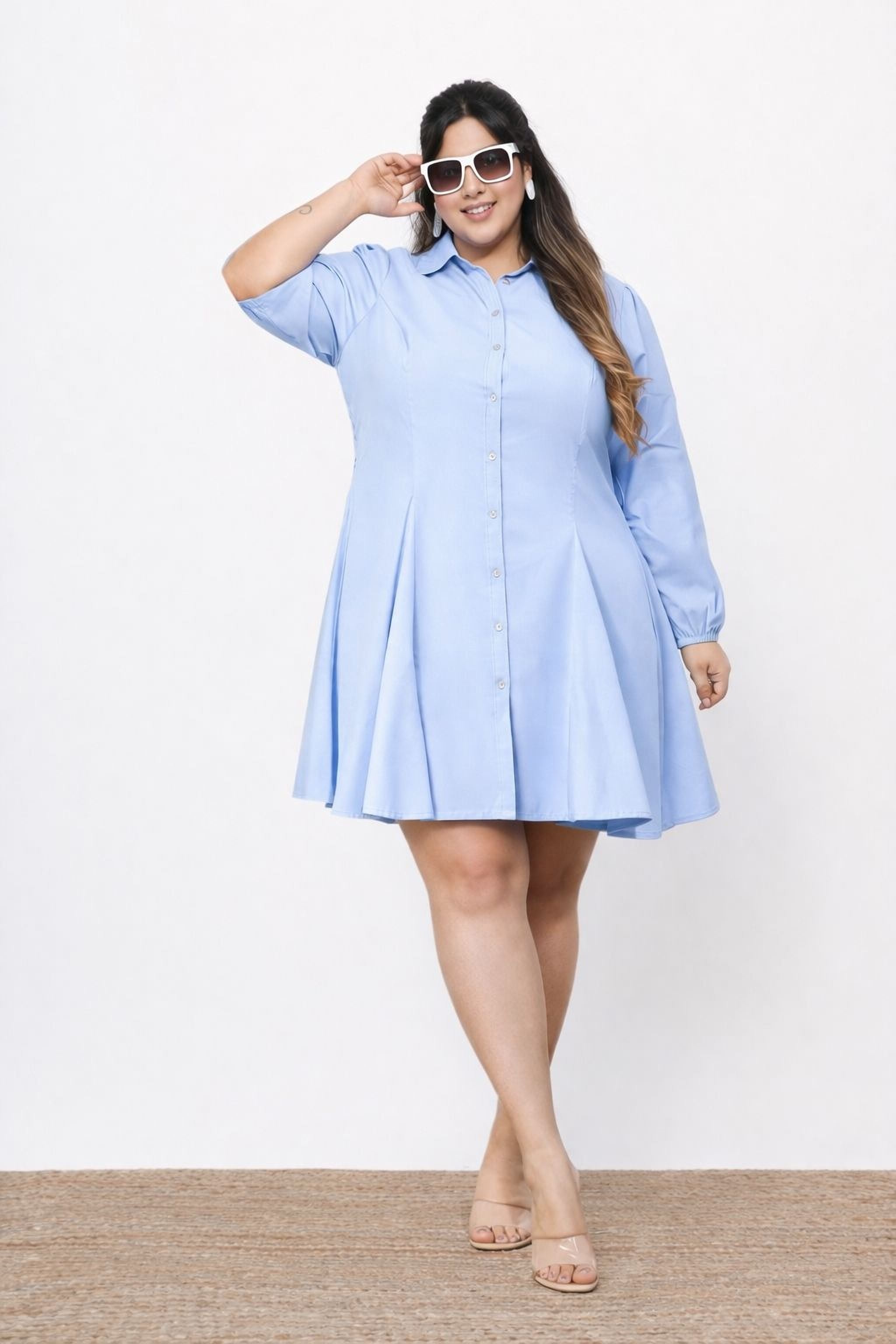 A woman wearing a light blue dress and sunglasses stands confidently against a white background.