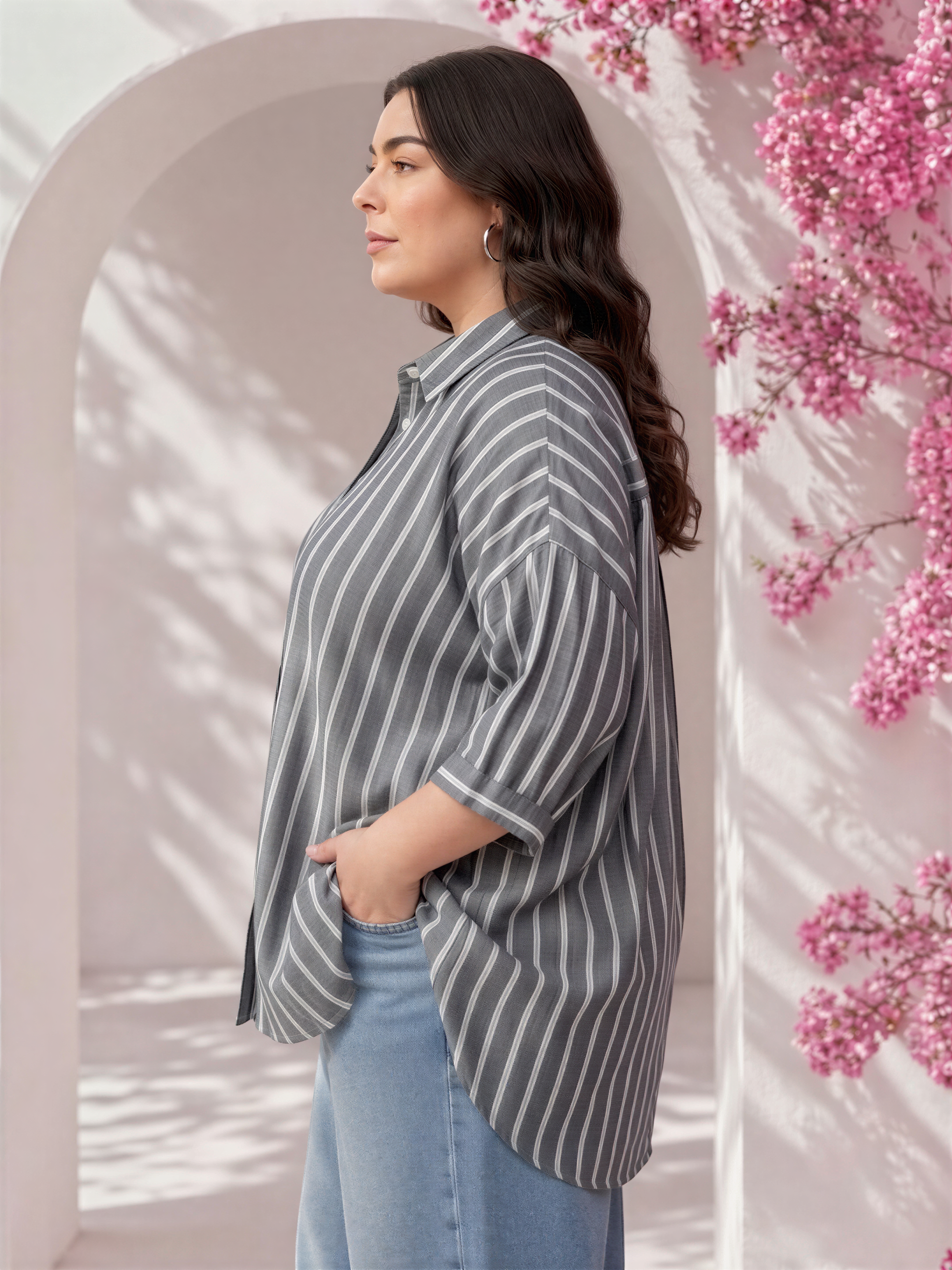 A woman wearing a striped shirt and jeans stands in front of a pink flowering archway, with her hands in her pockets.