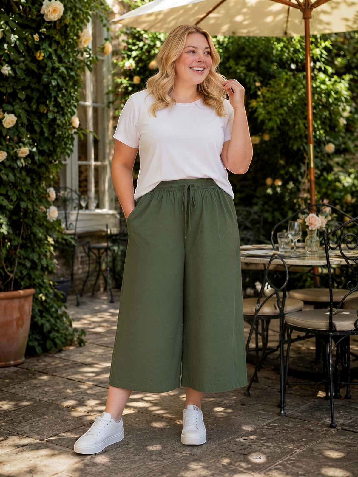 A woman wearing a white t-shirt, olive green wide-leg pants, and white sneakers stands in a garden setting with a table and chairs nearby.
