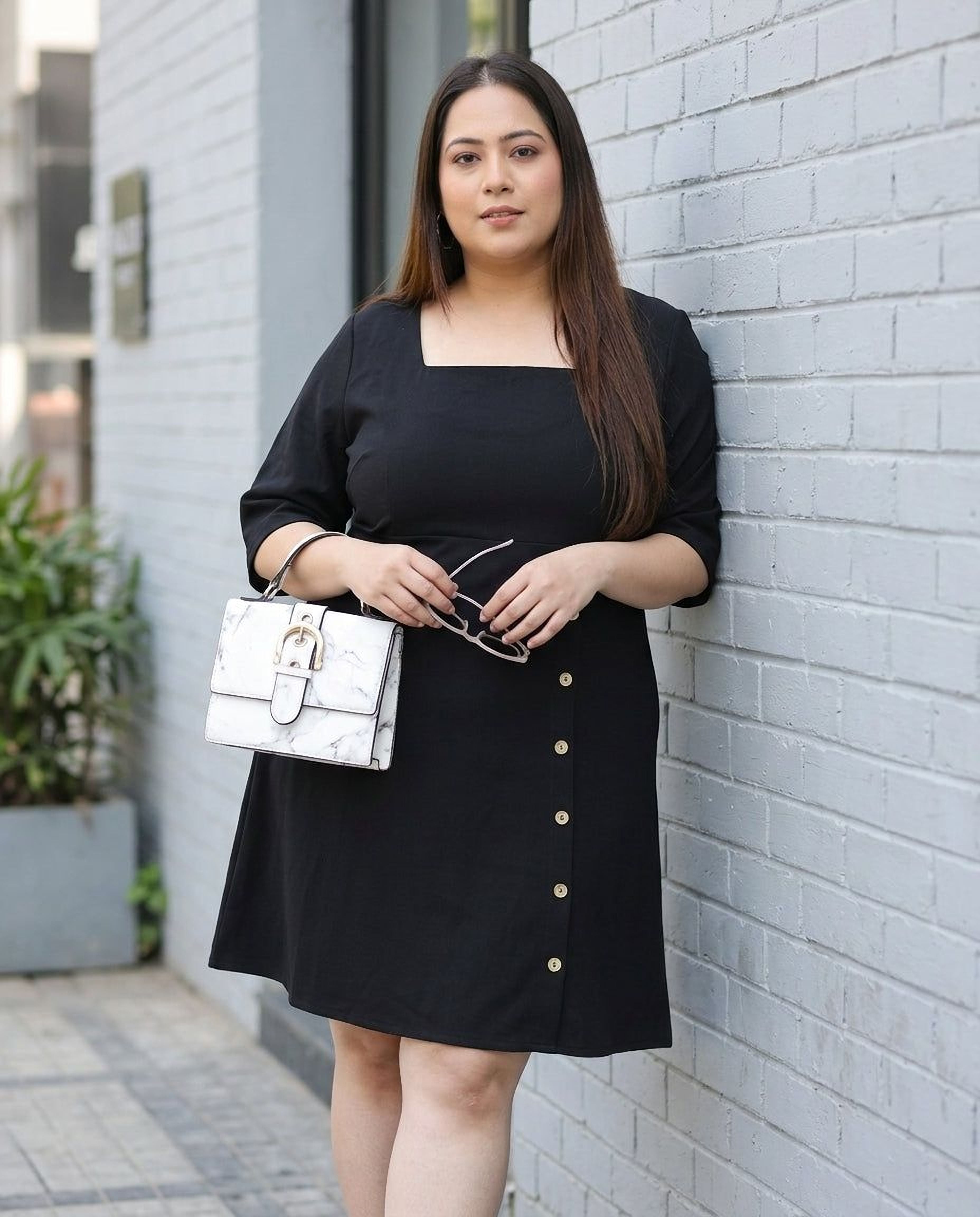 Woman in plus size black dress with square neckline and side buttons, holding a white handbag