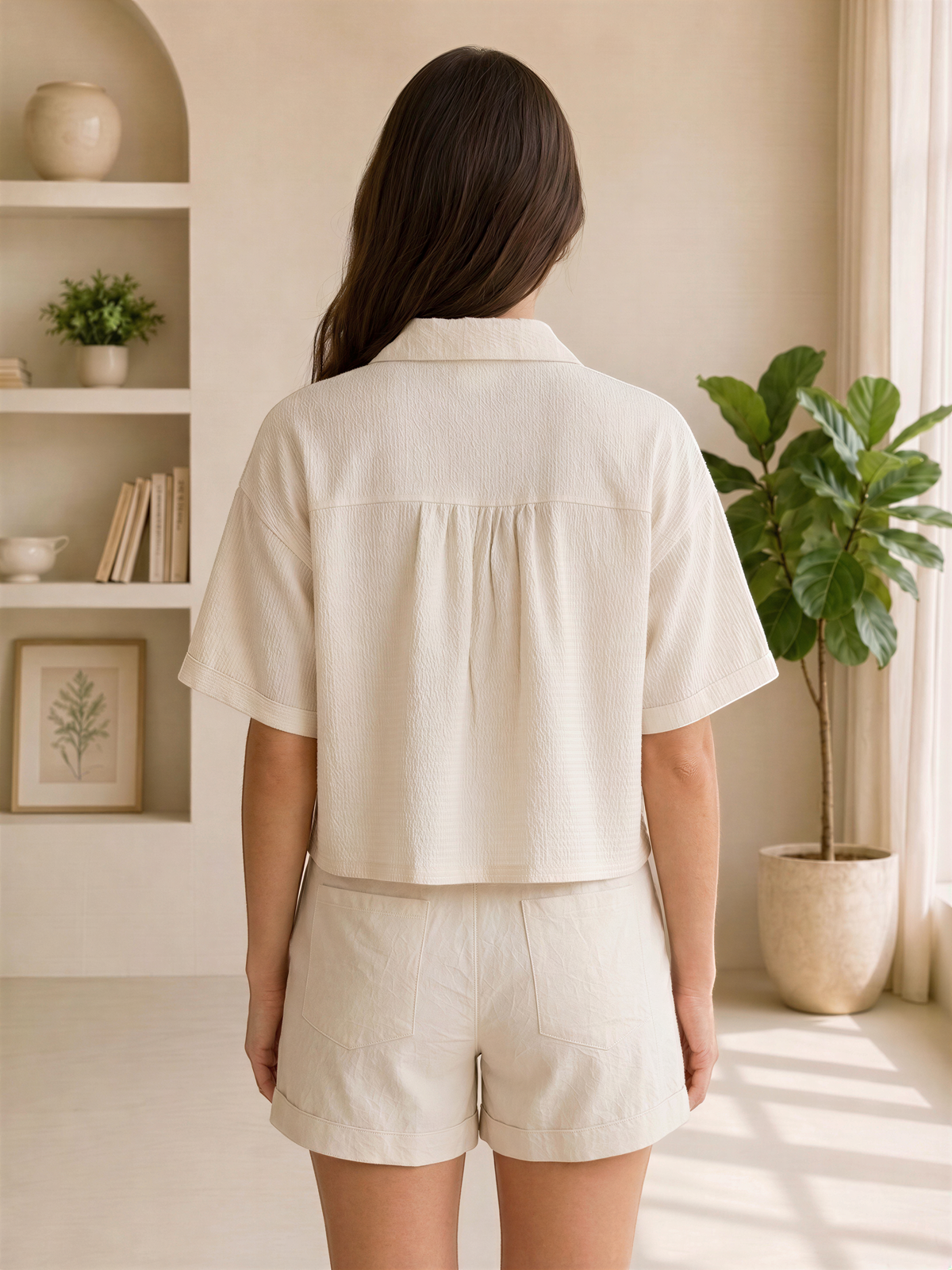 A woman wearing a white shirt and shorts stands in a room with a window, plants, and shelves.