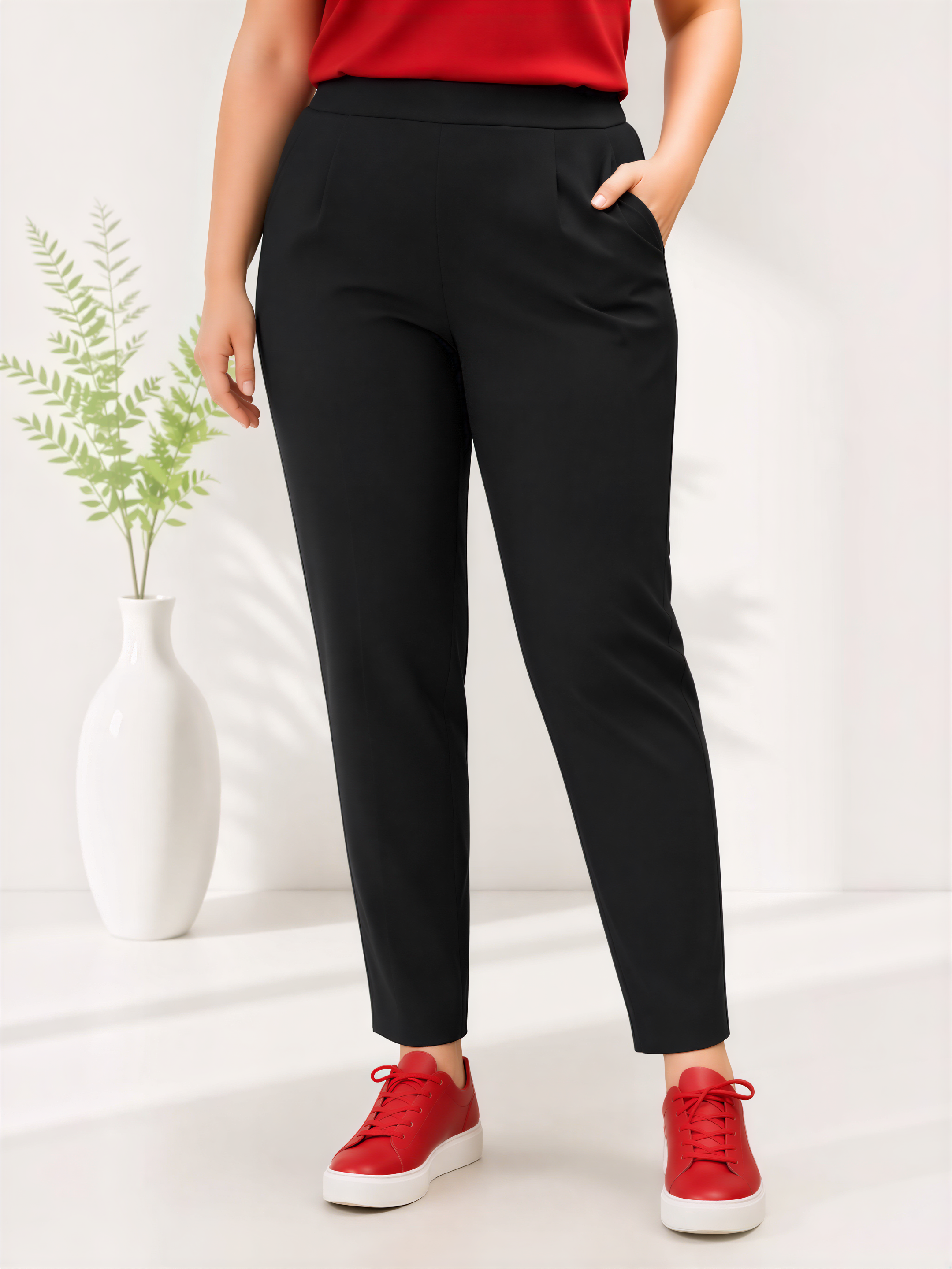 A person wearing a red top and black pants stands in front of a white vase with a plant, wearing red shoes.