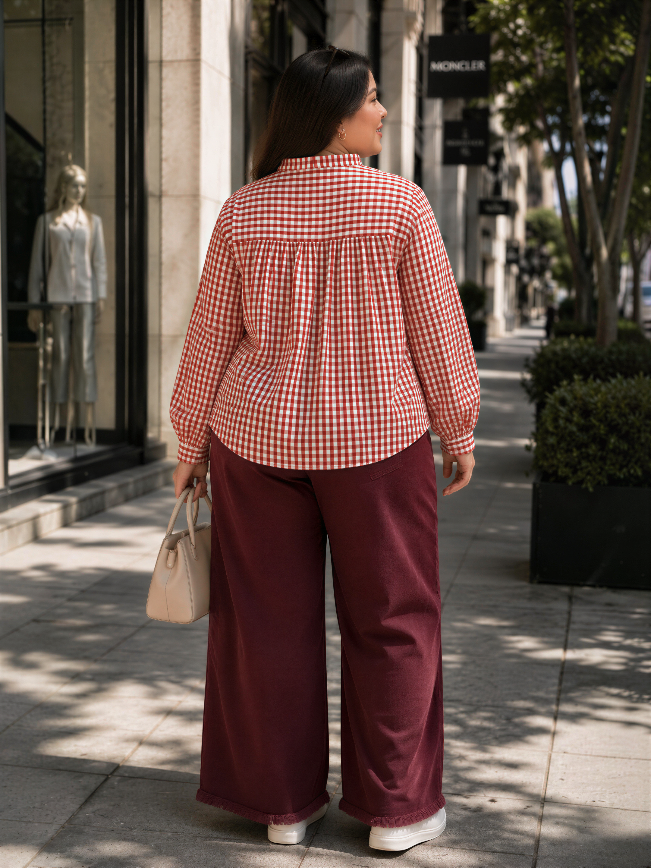 A woman wearing a red and white checkered shirt and maroon wide-leg pants is standing on a sidewalk, looking away from the camera.