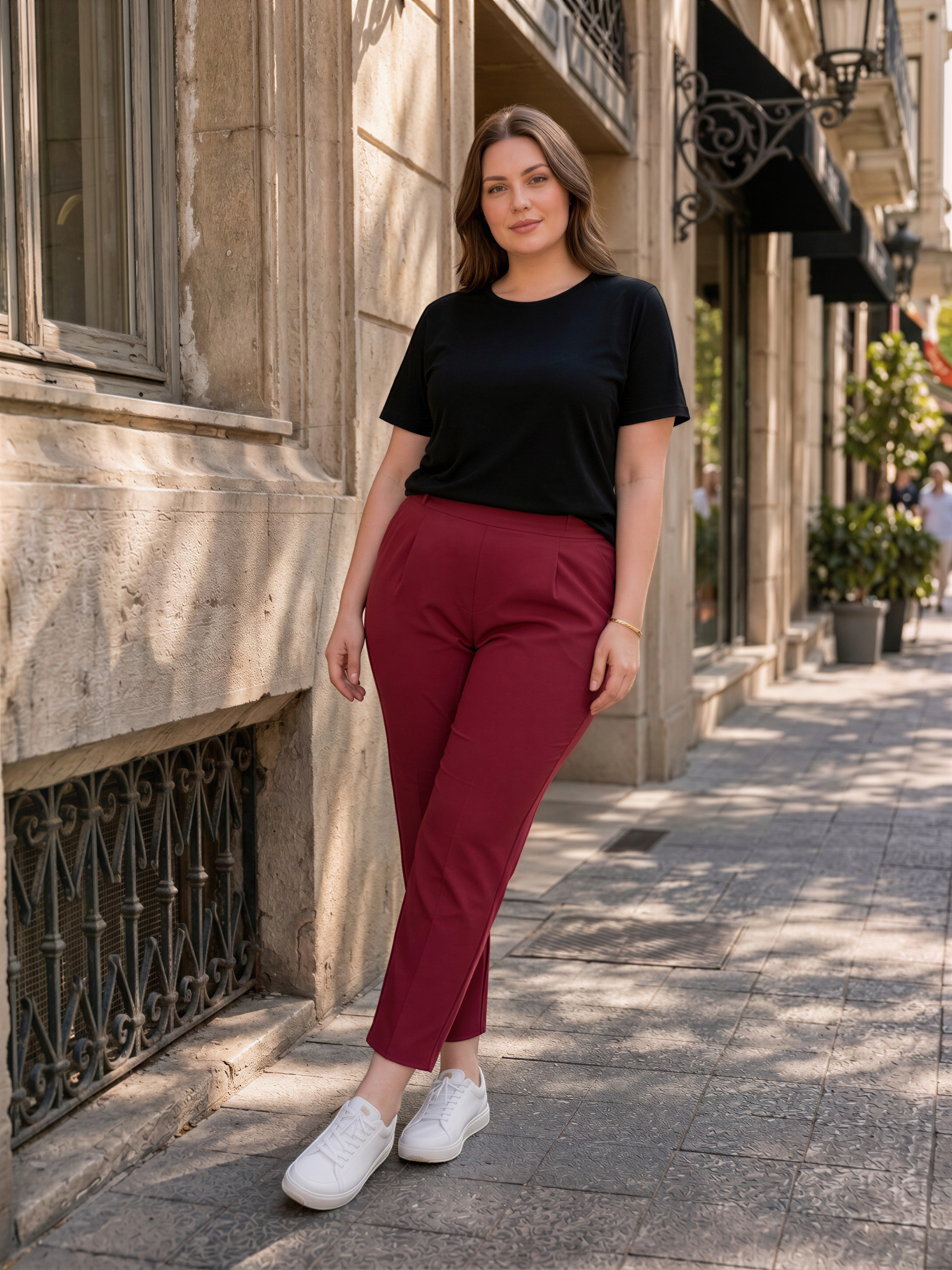 A woman wearing a black top and red pants stands on a sidewalk, posing for the camera.