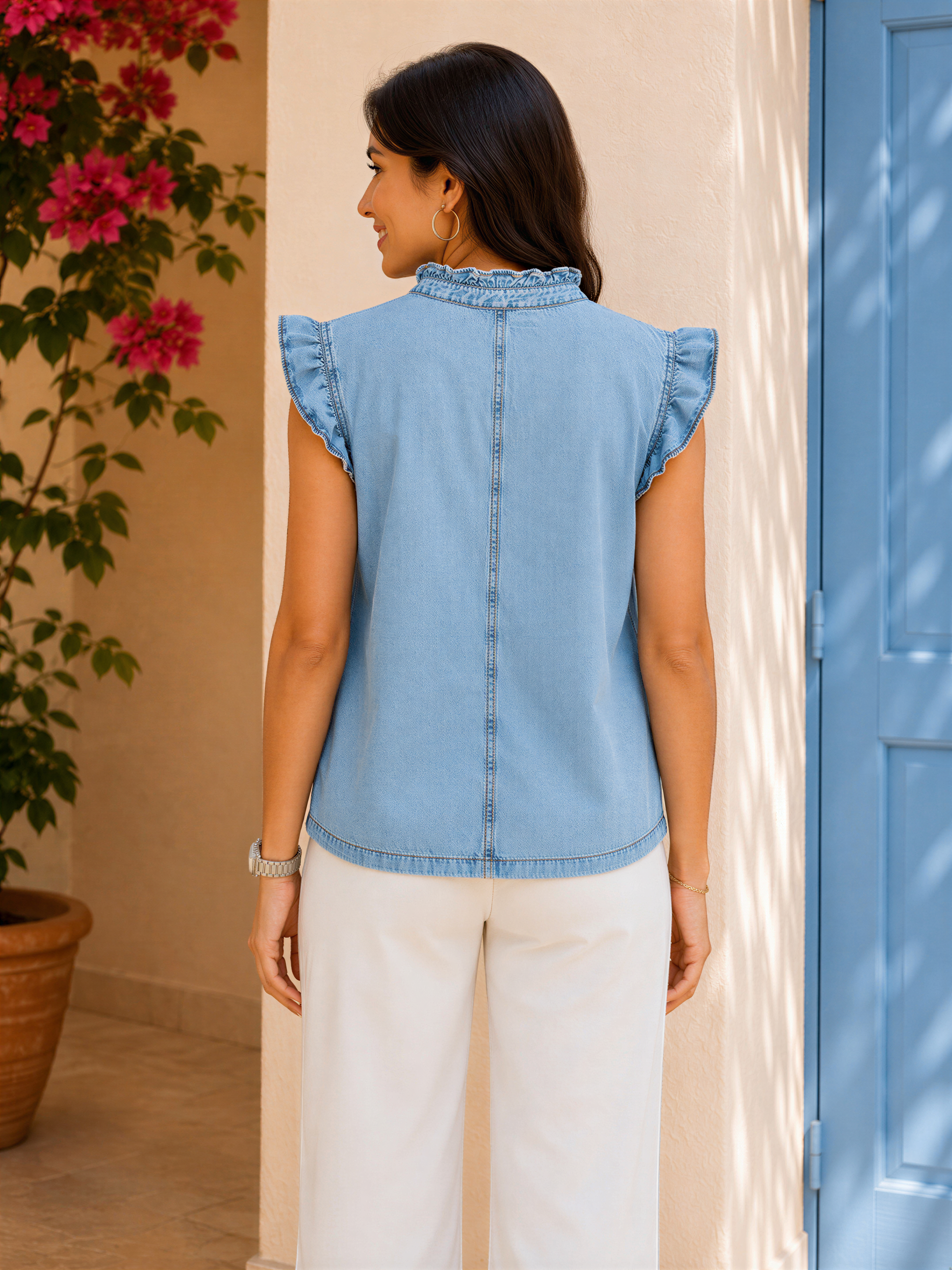 A woman wearing a blue denim sleeveless top and white wide-leg pants stands in front of a blue door and pink flowers.