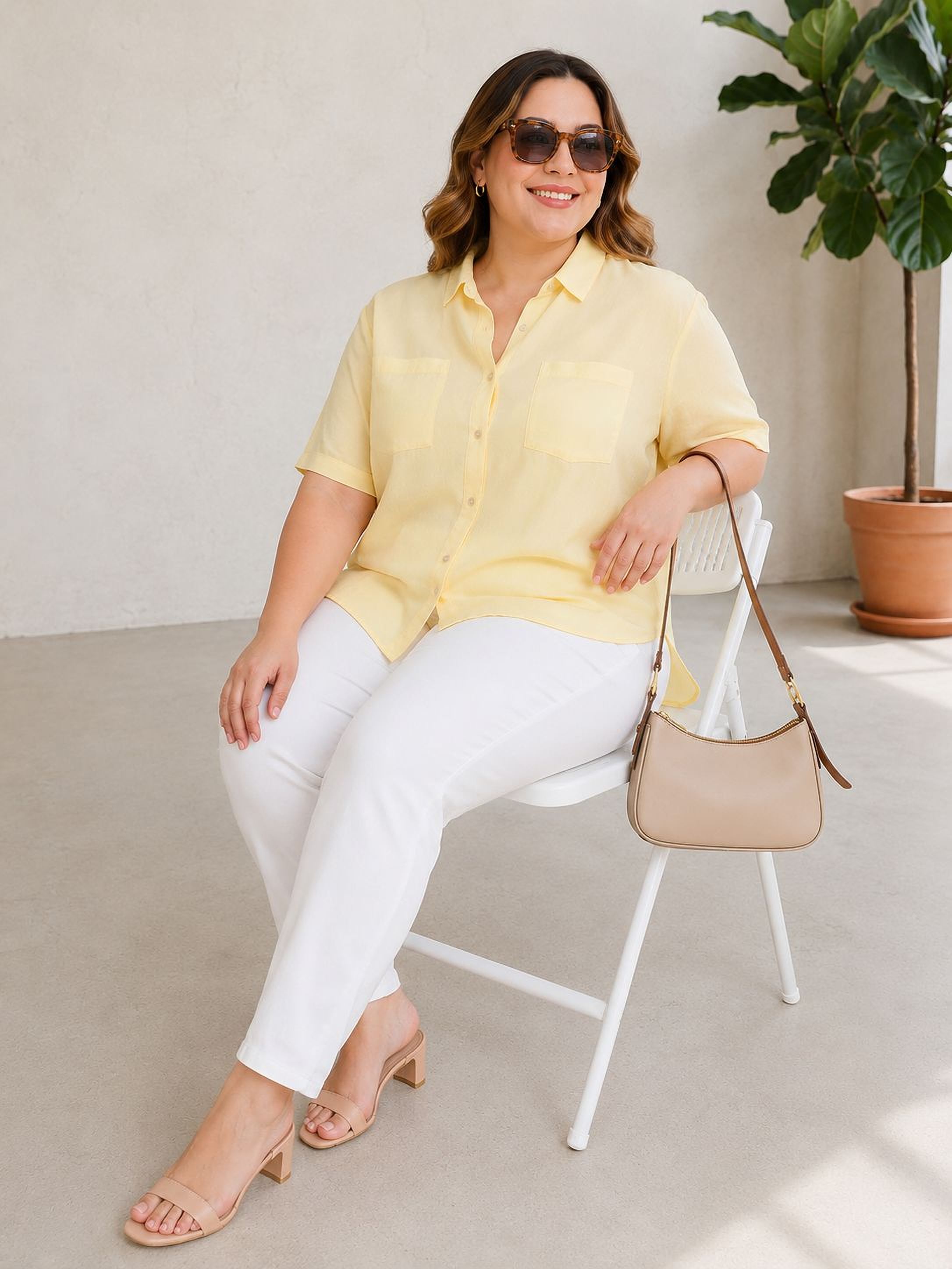 A woman wearing a yellow blouse and white pants is sitting on a white folding chair, smiling and holding a beige purse.