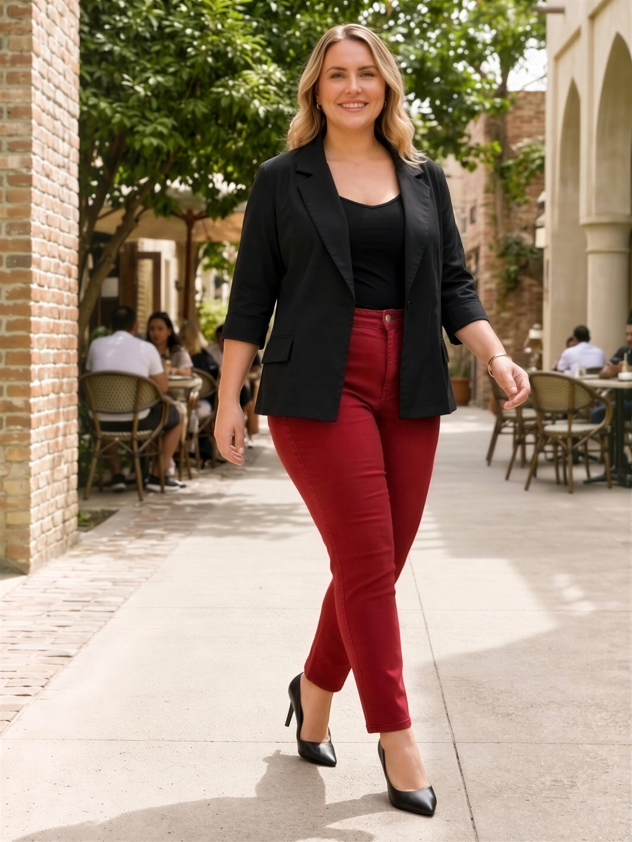 A woman wearing a black blazer, red pants, and black heels is walking down a sidewalk, smiling and looking at the camera.