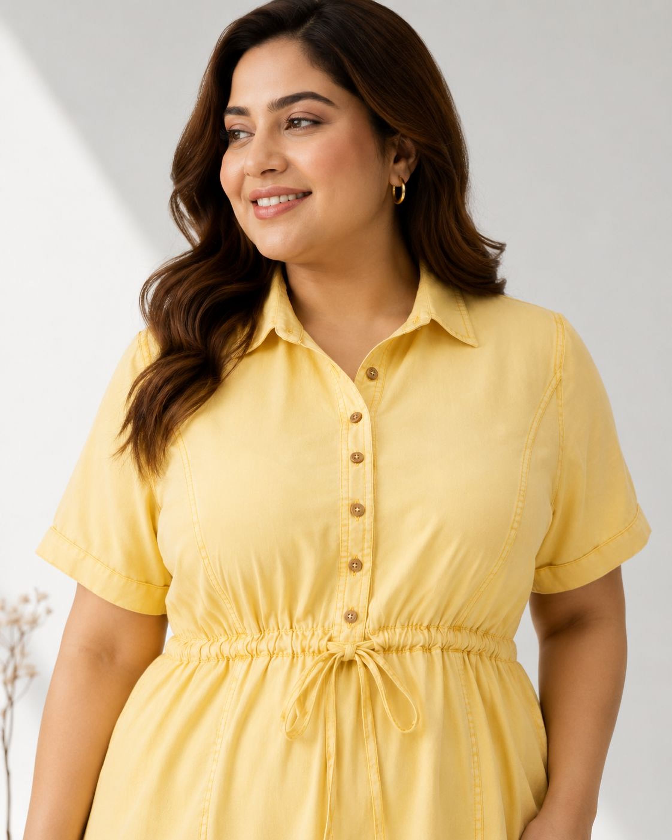 A woman with long brown hair wearing a yellow dress with a ruffled waist and buttons, smiling at the camera.