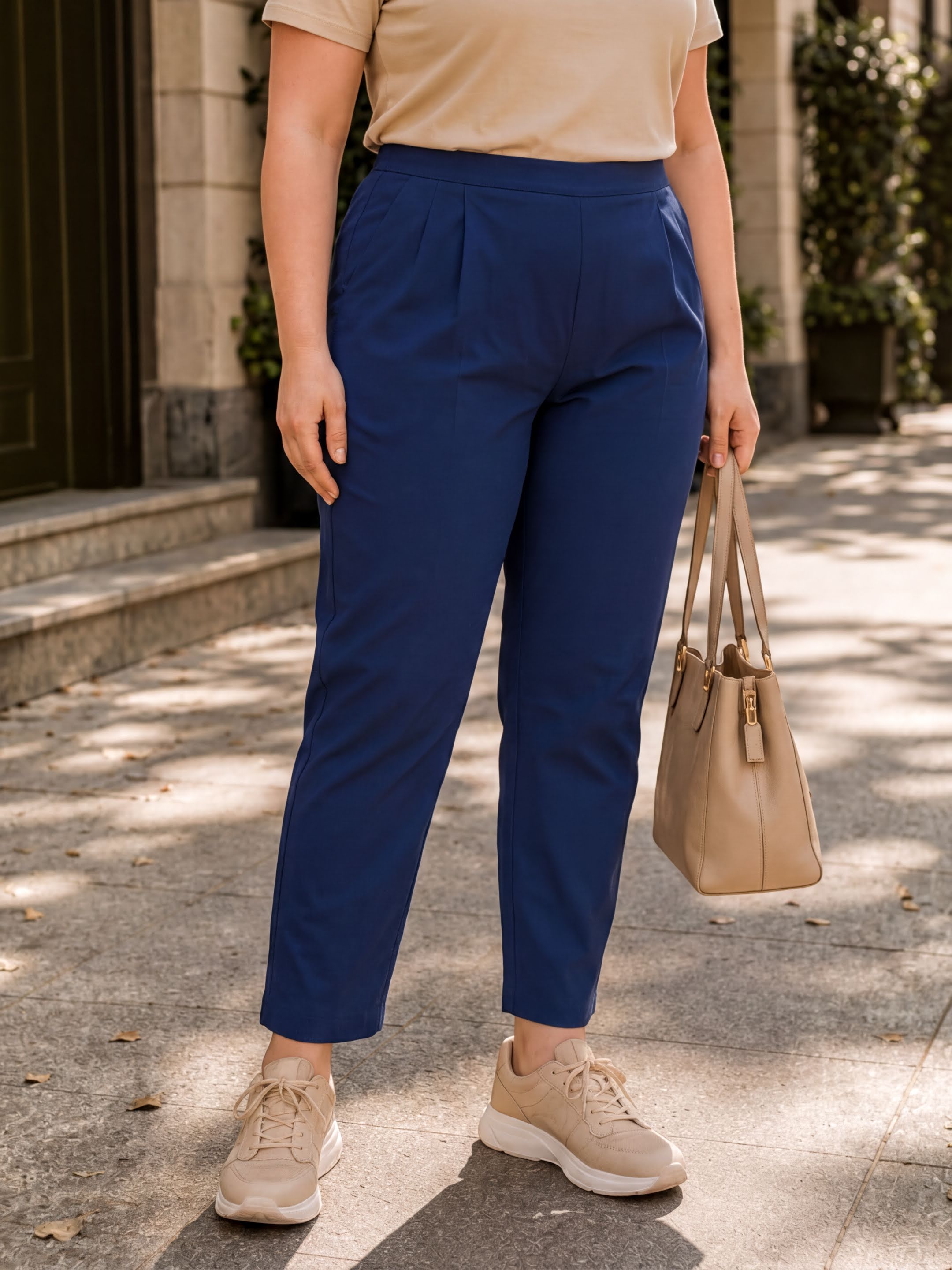 A woman wearing blue pants and beige shoes is standing on a sidewalk, holding a beige handbag.