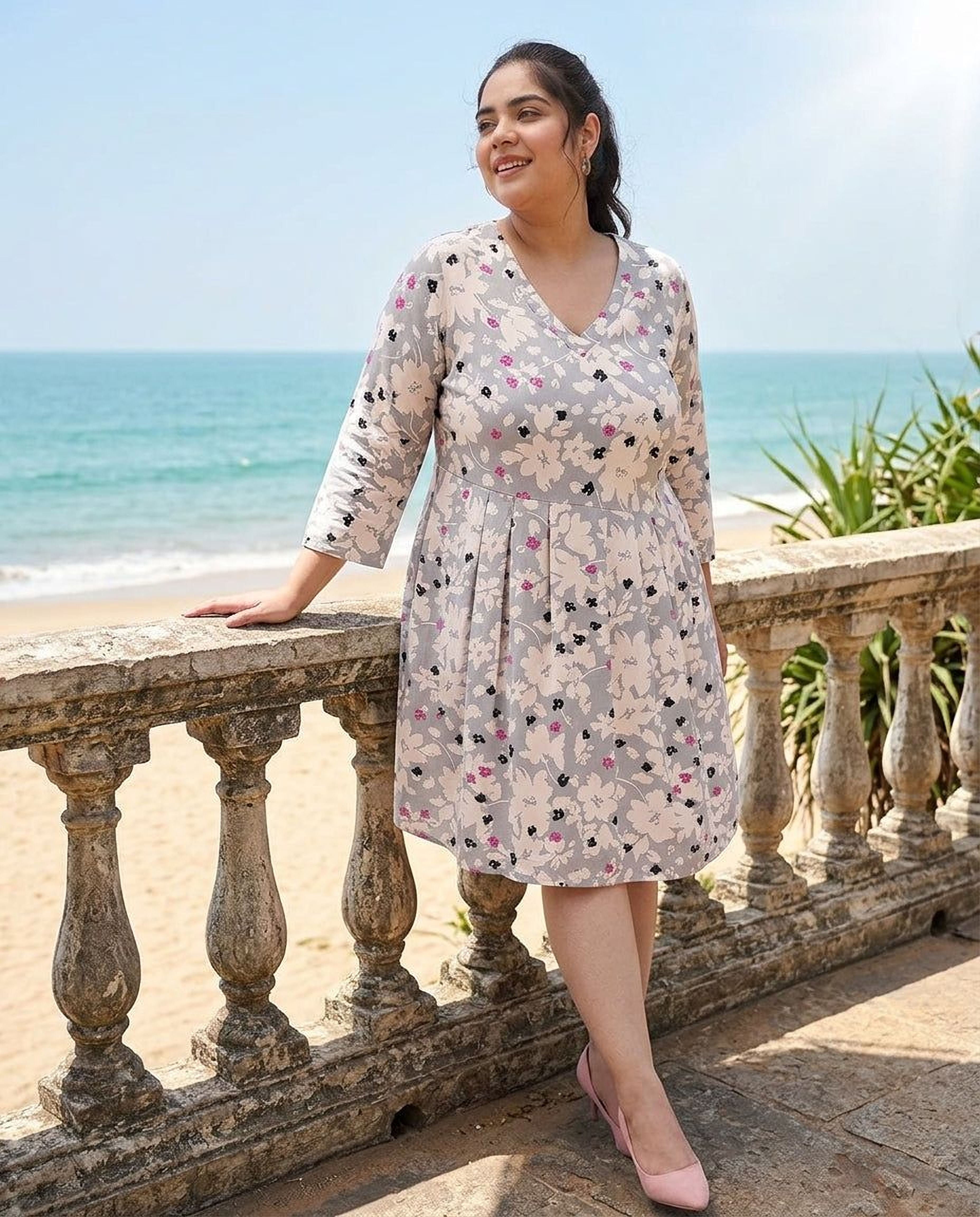 A woman wearing a gray and white floral print dress with a v-neck and long sleeves, smiling and posing with her hand on her face.