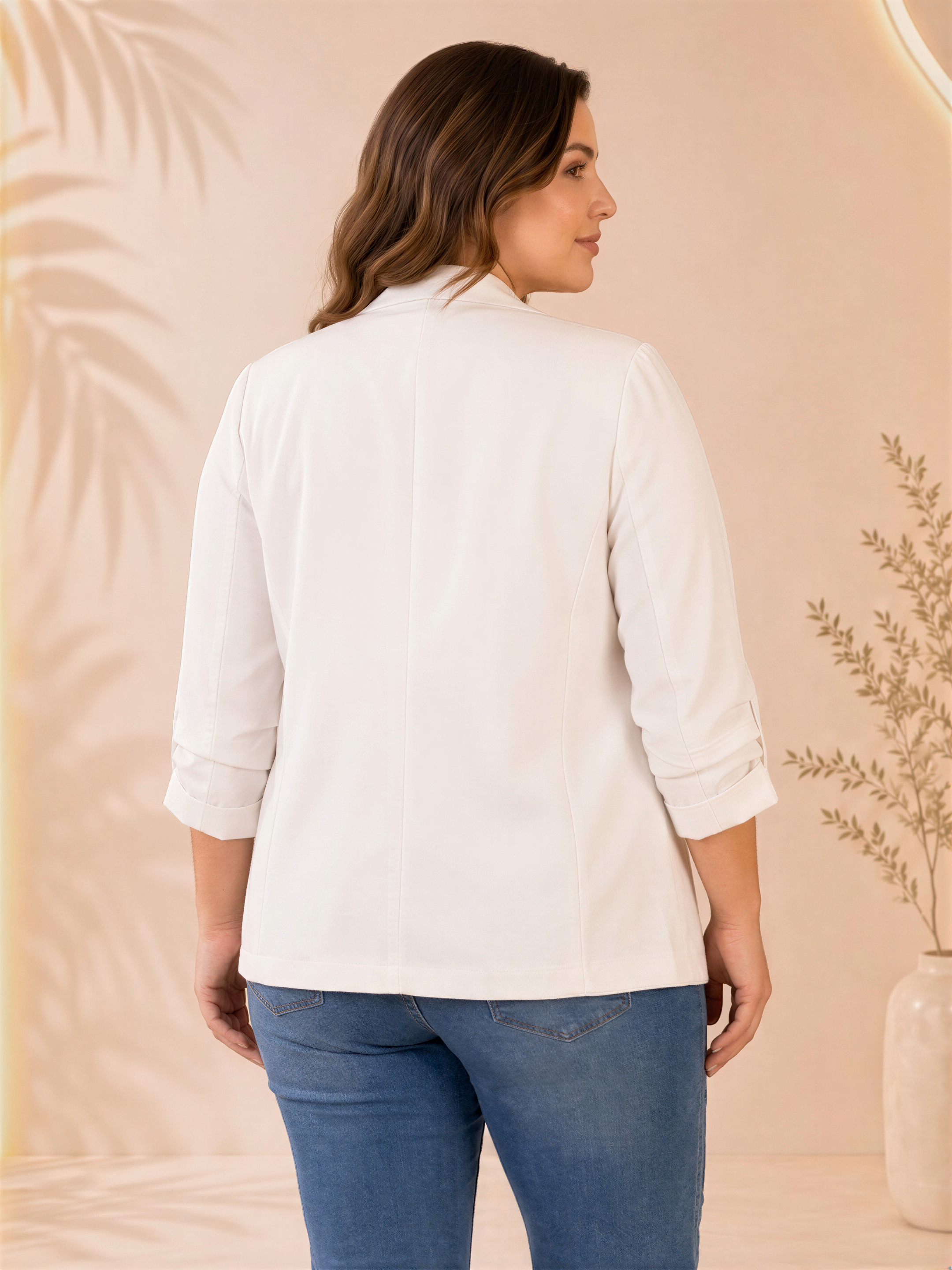 A woman wearing a white blazer and blue jeans stands in front of a peach-colored background with palm leaves.