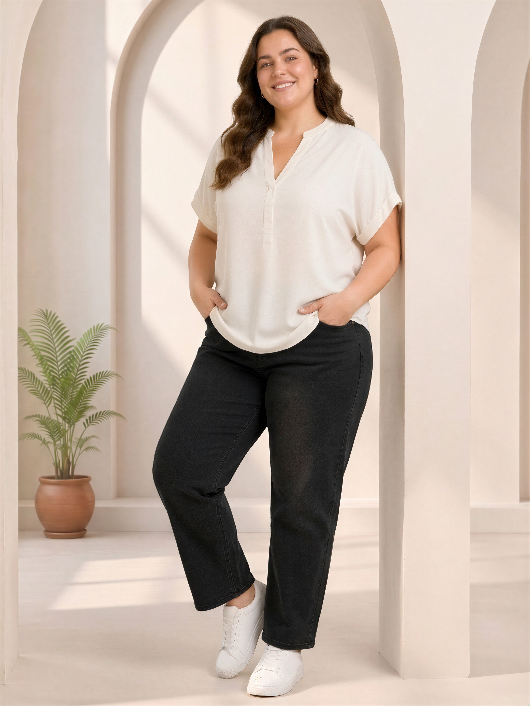 A woman in a white blouse and black pants stands in a room with arched doorways and a potted plant.