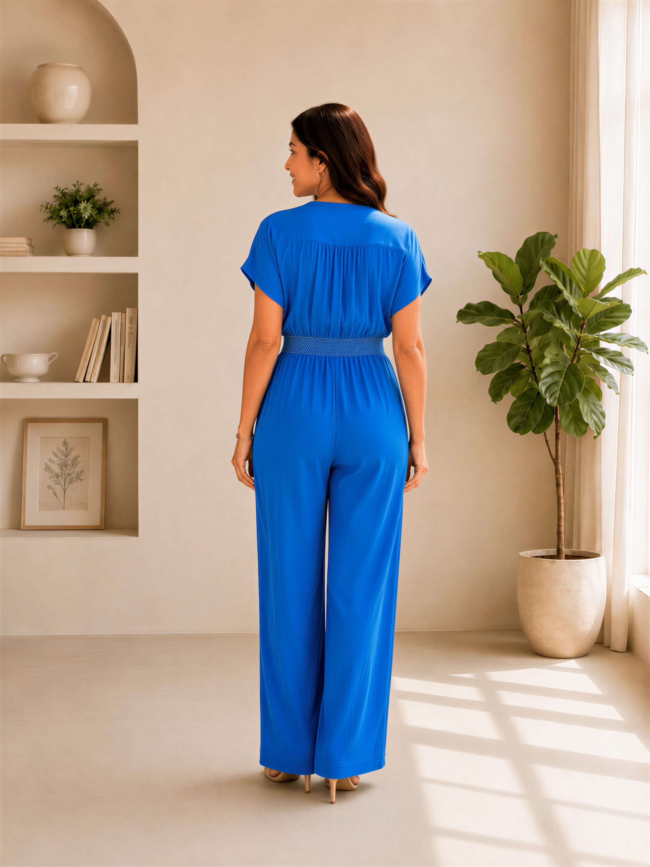 A woman in a blue jumpsuit stands in a room with a window, bookshelf, and potted plant.