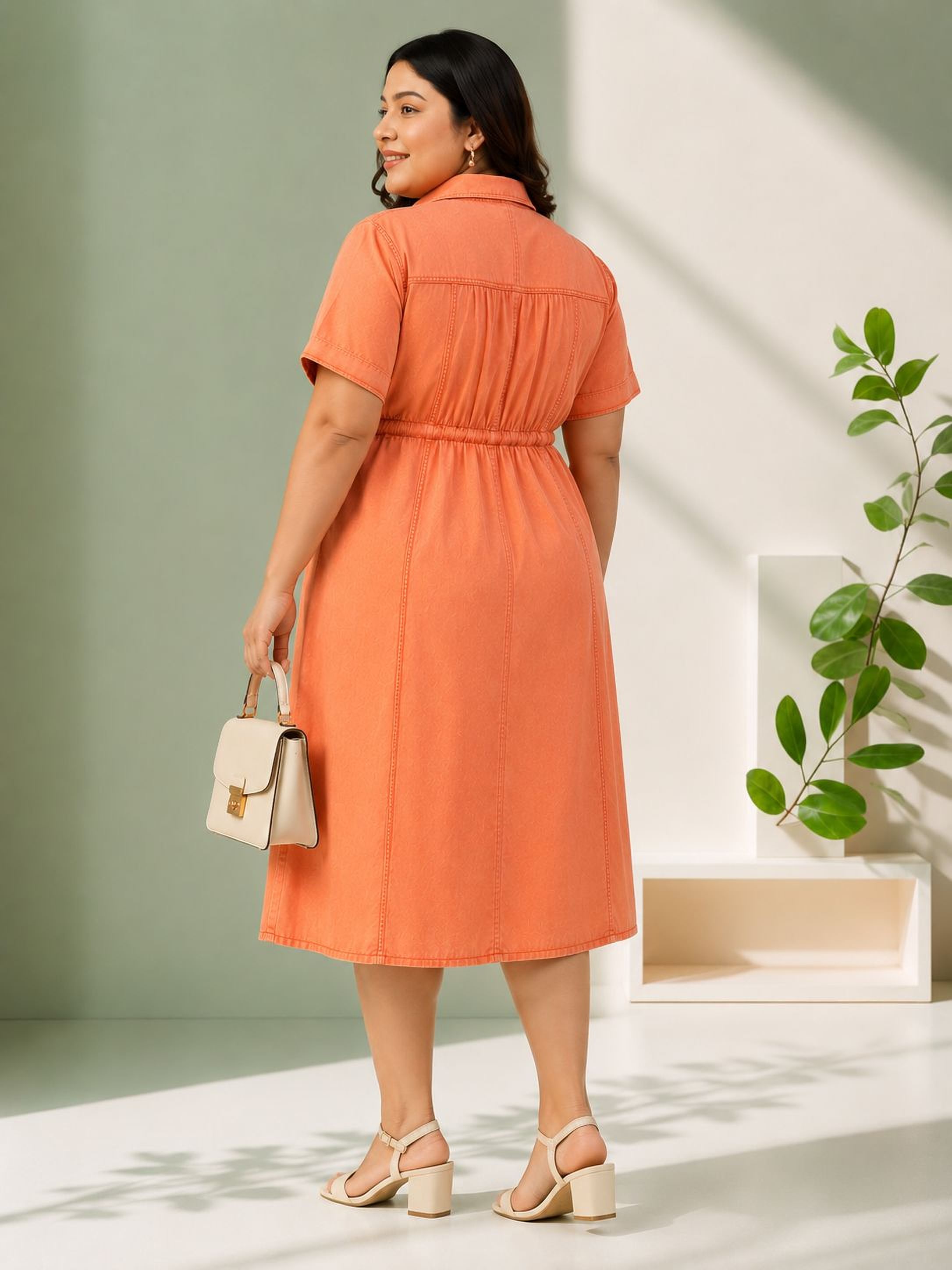 A woman wearing an orange dress stands in a minimalist setting, with a potted plant and a white shelf in the background.