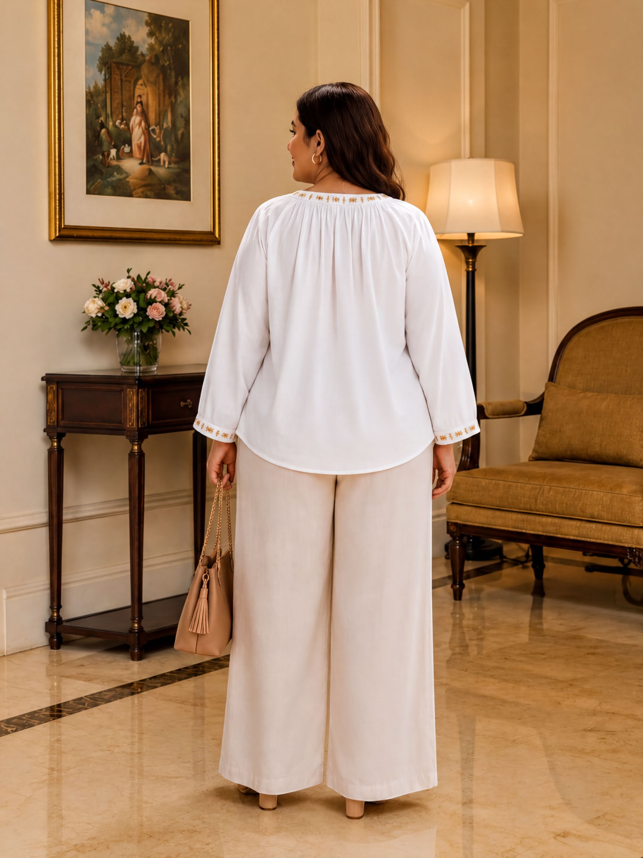 A woman in a white blouse and wide-leg pants stands in a room with a painting on the wall, a table with flowers, and a lamp.