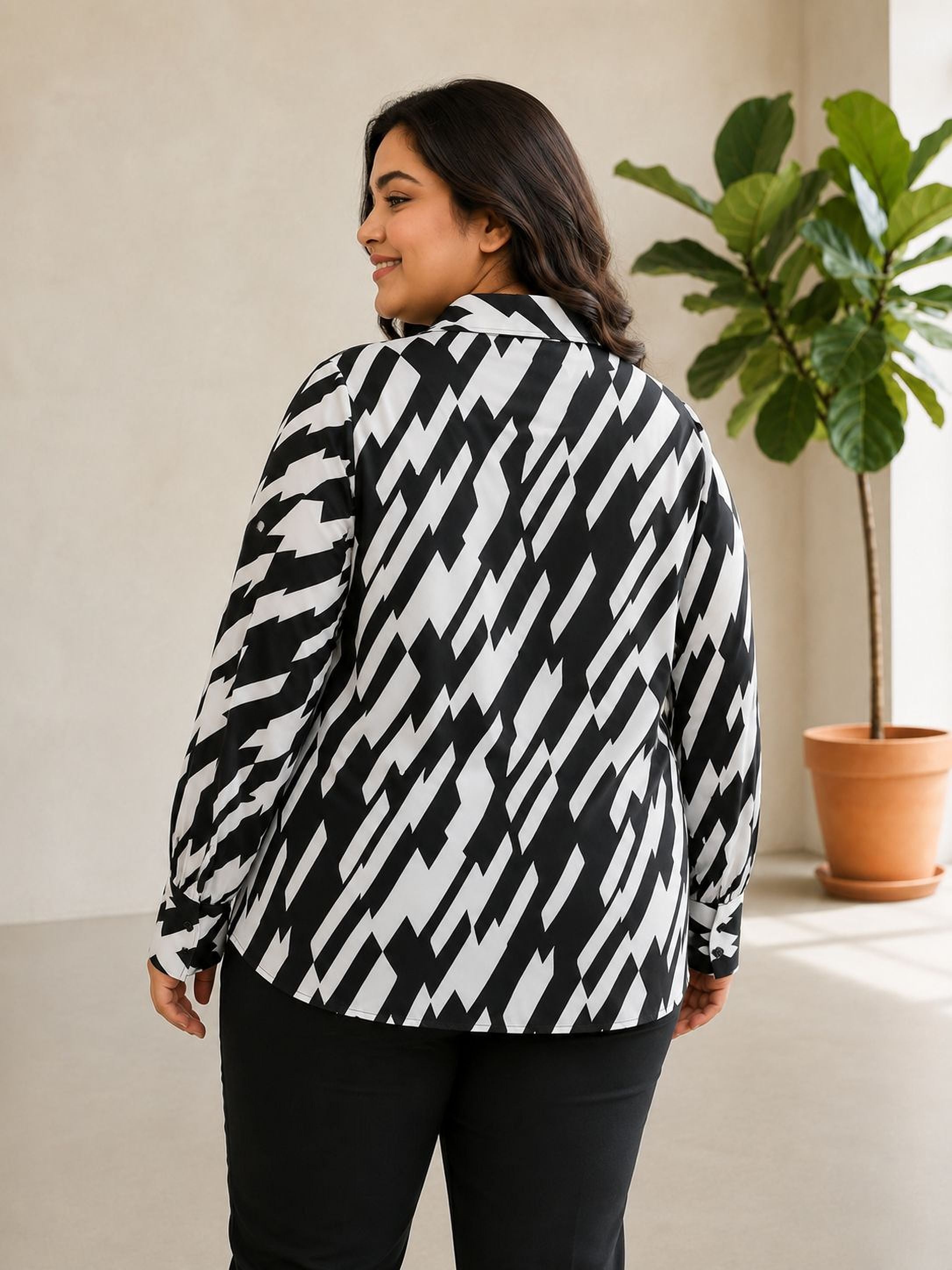 A woman wearing a black and white patterned blouse stands in a room with a potted plant in the background.