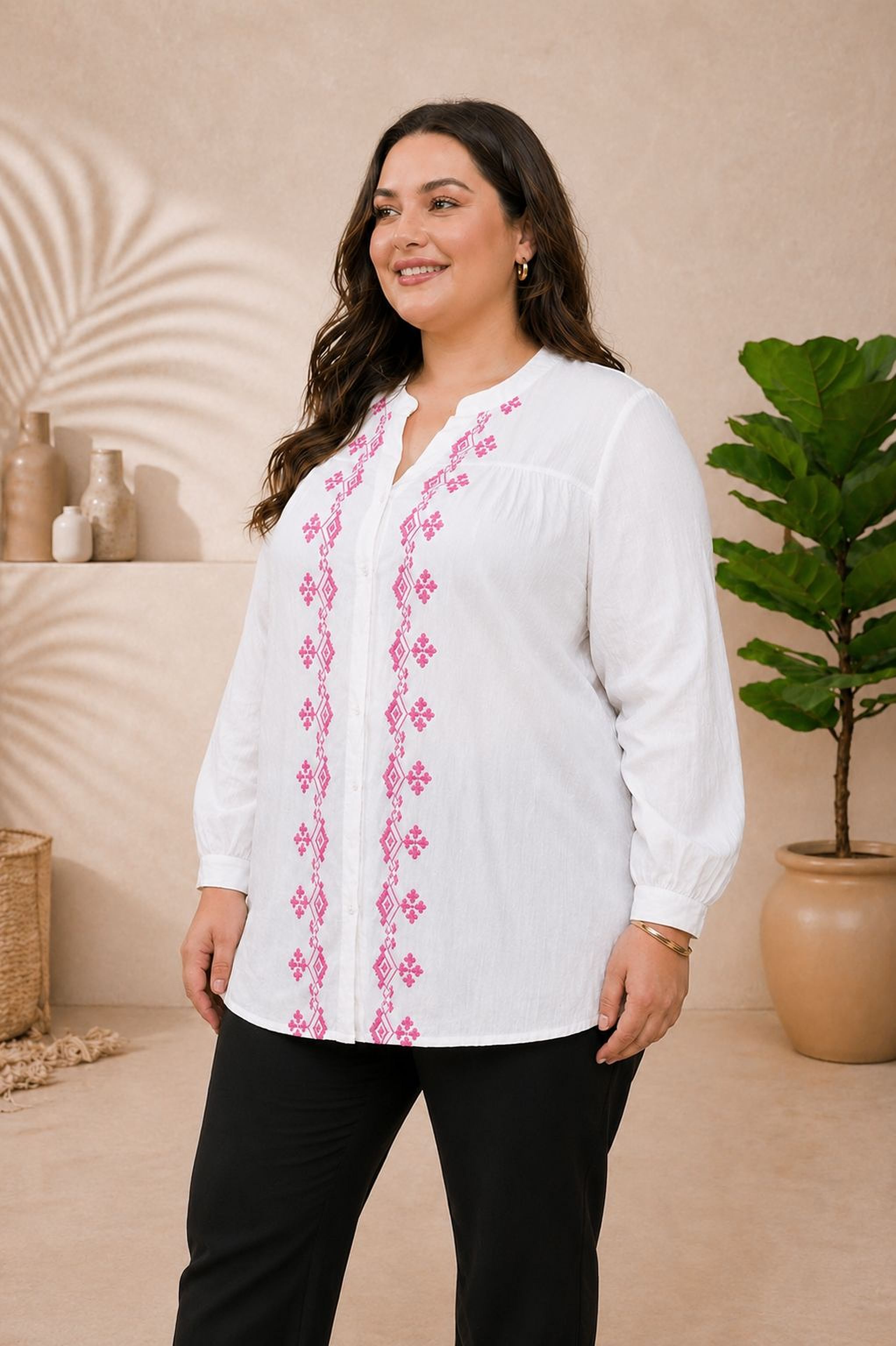 A woman wearing a white blouse with pink embroidered designs stands in a room with a potted plant and a shelf with decorative items.
