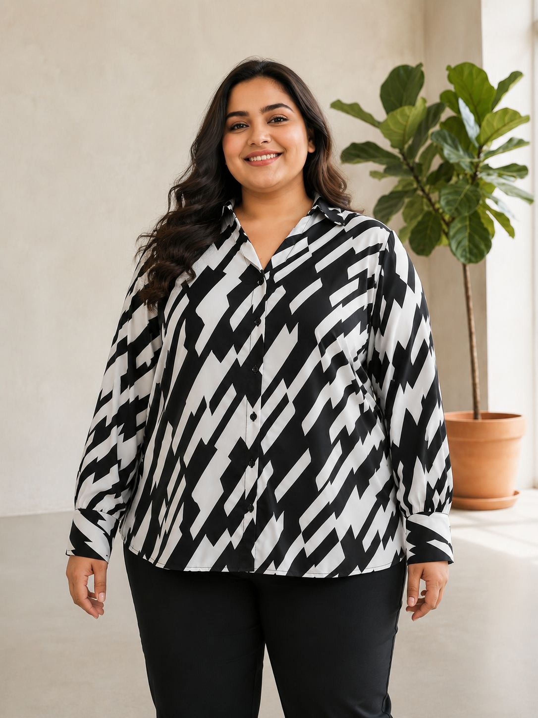 A woman with long dark hair is standing in a room, wearing a black and white patterned blouse and black pants. She is smiling and looking directly at the camera.