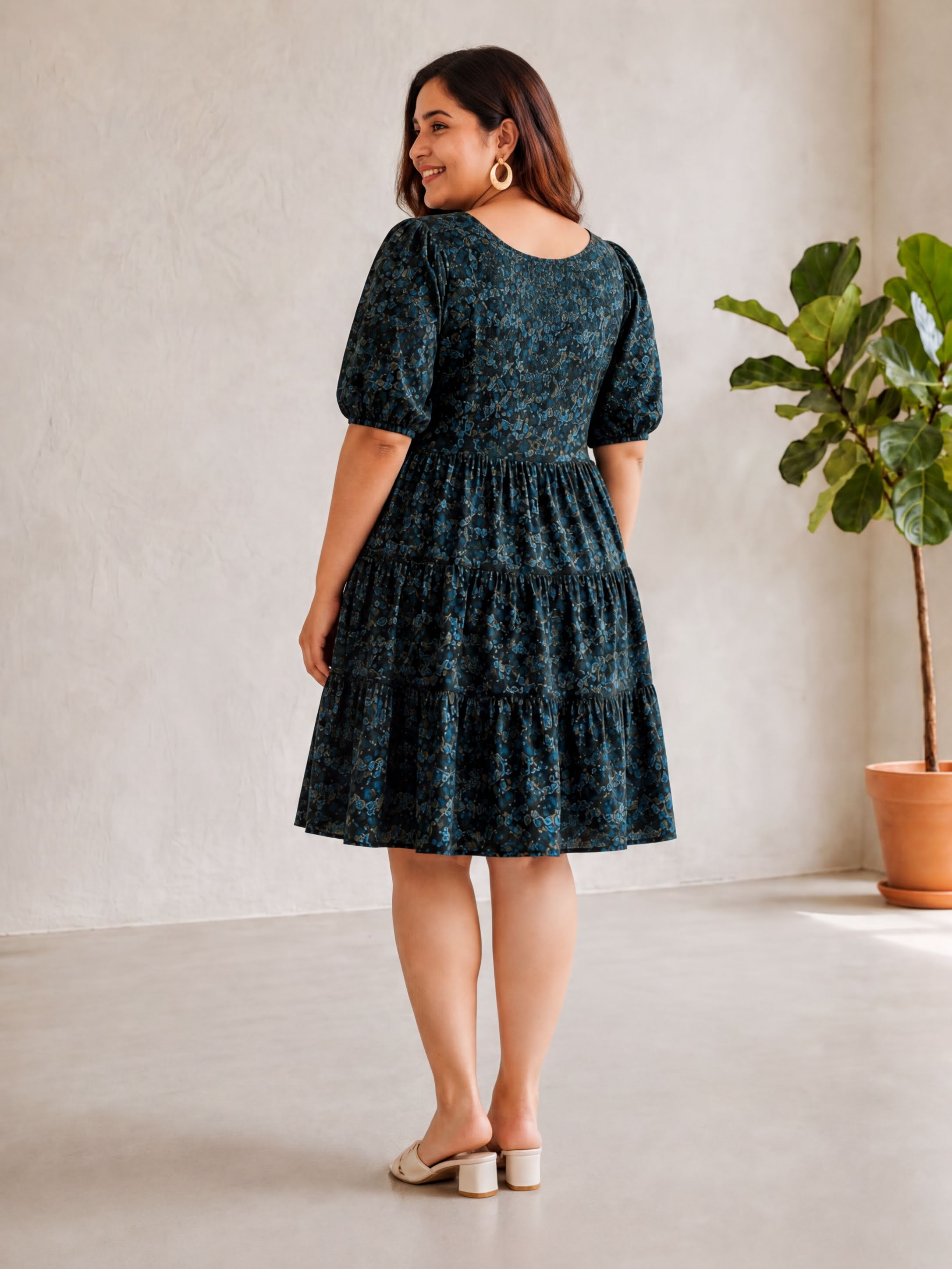 A woman wearing a green floral print dress stands in a room with a potted plant.