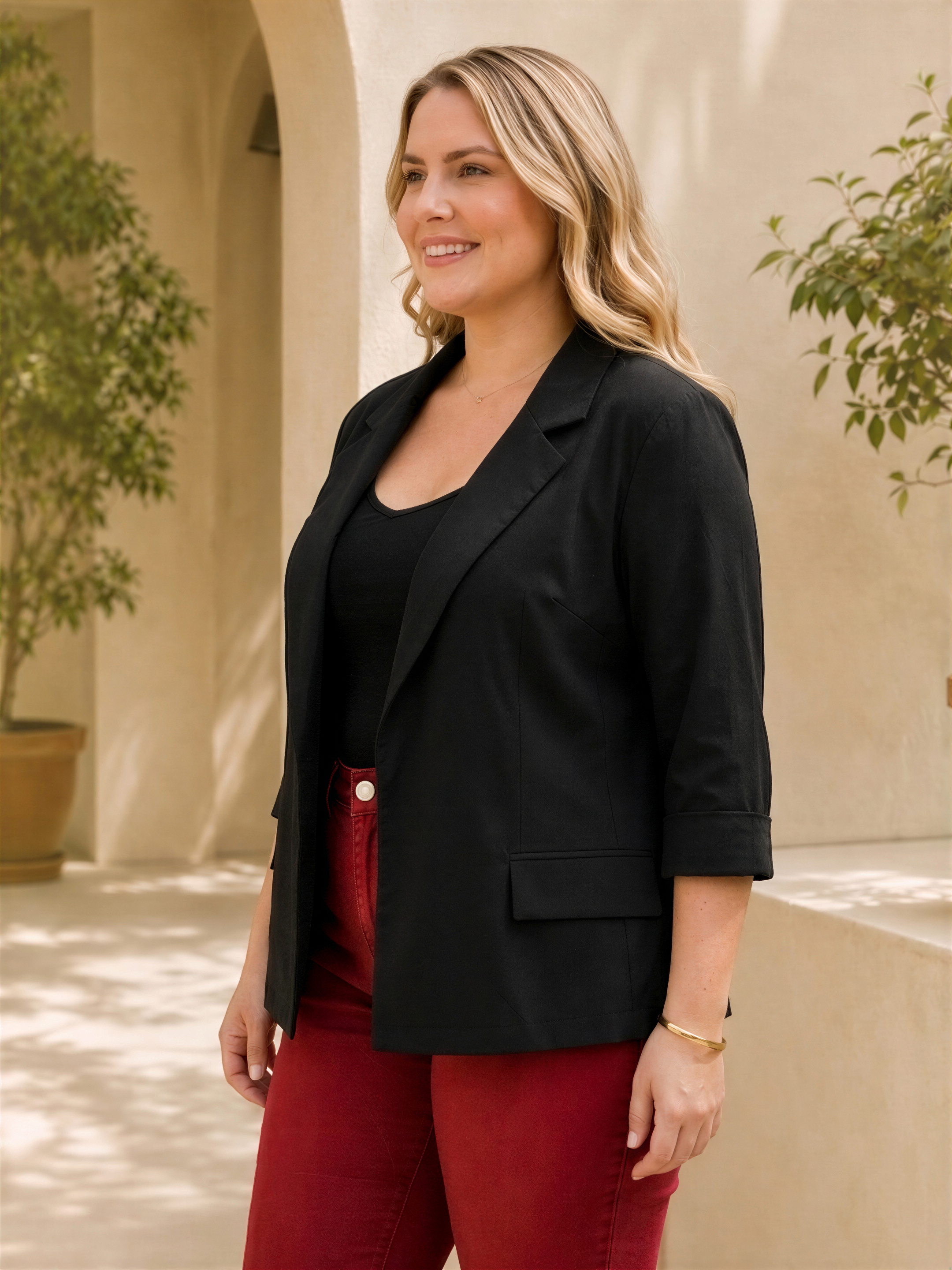 A woman with blonde hair is standing in front of a building, wearing a black blazer and red pants.
