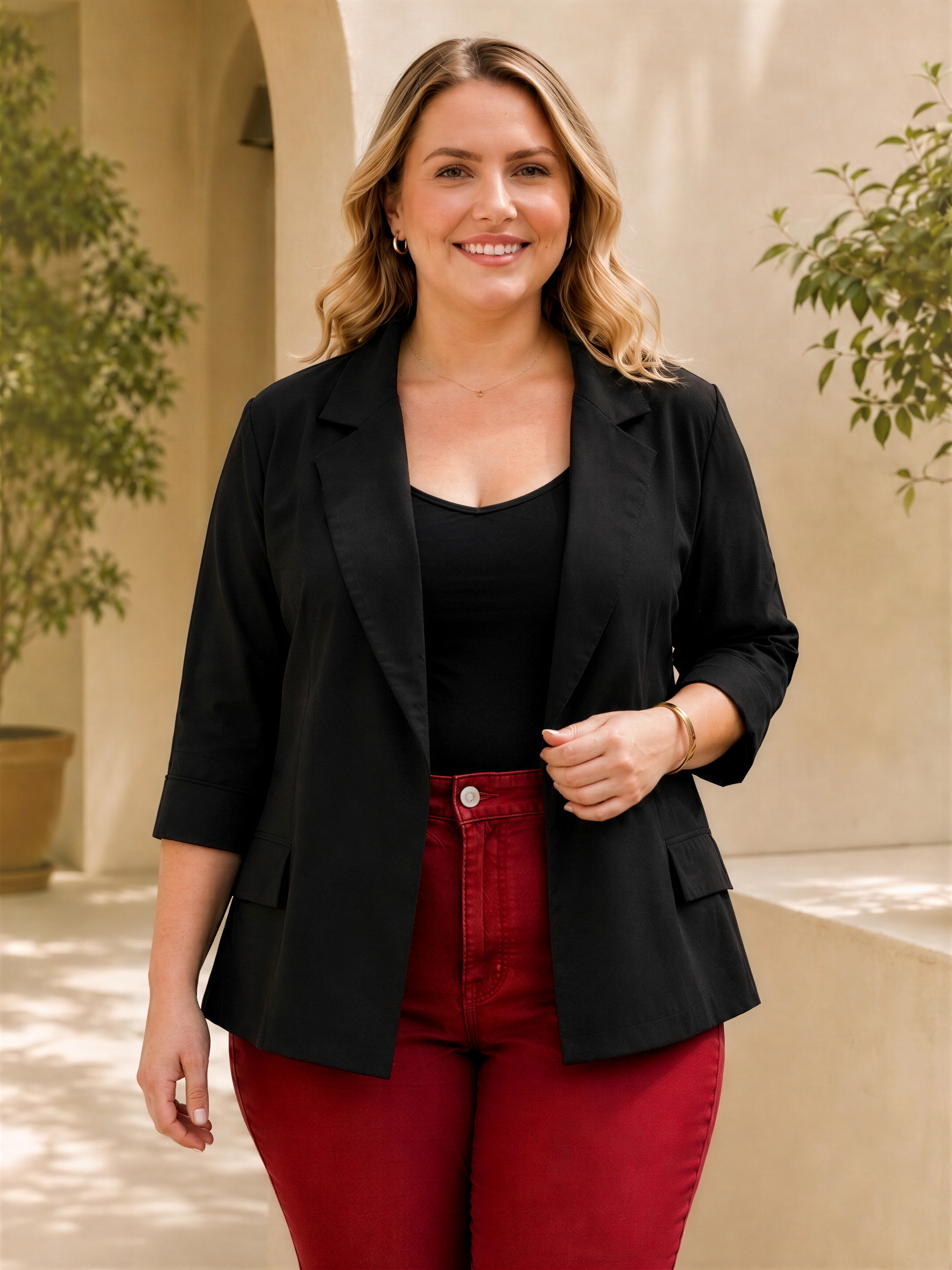 A woman with blonde hair is standing in front of a building, wearing a black blazer, a black top, and red pants. She is smiling and appears to be posing for the camera.