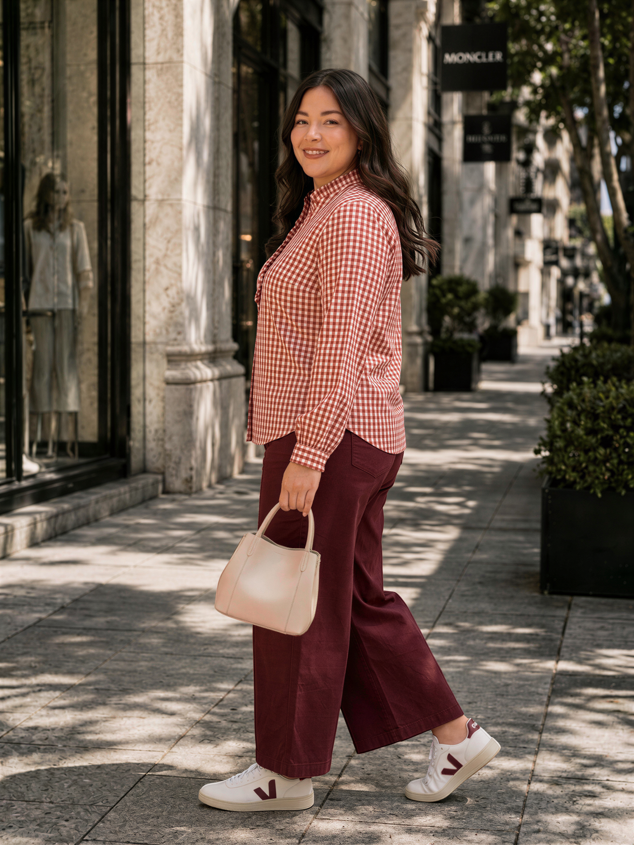 A woman wearing a red and white checkered shirt, burgundy pants, and white sneakers is walking down a city sidewalk, carrying a white handbag.
