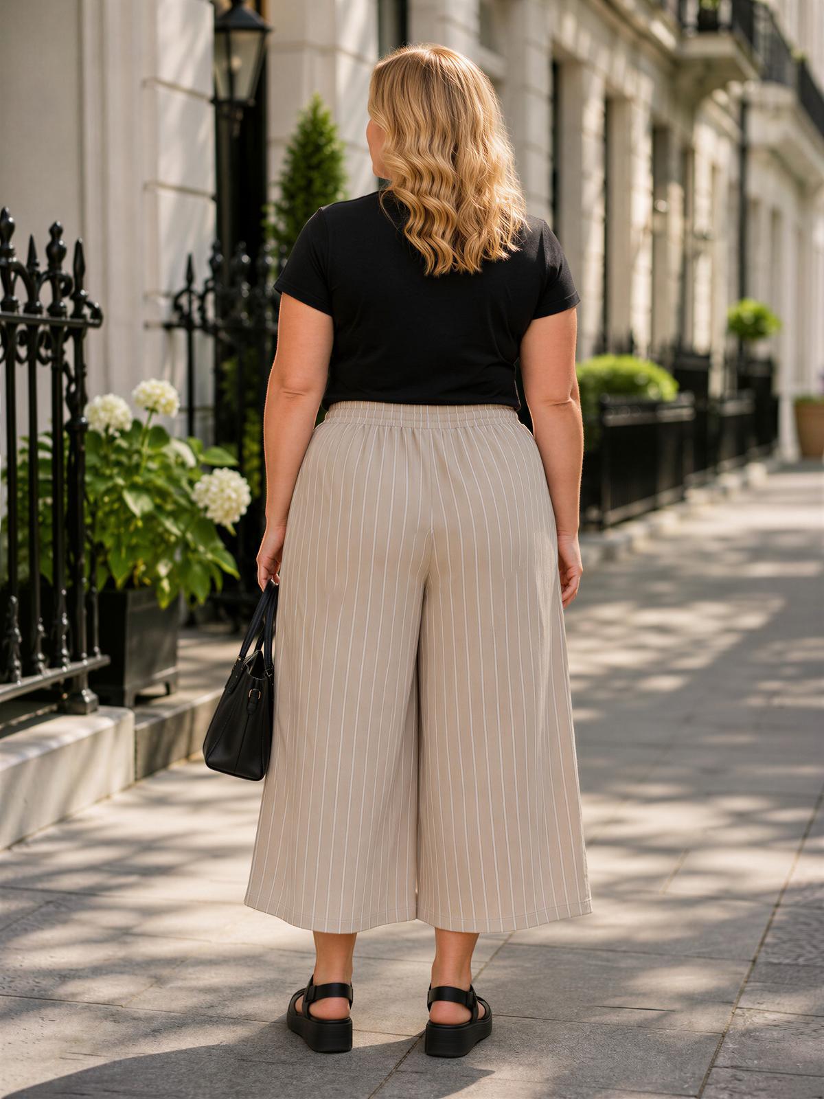 A woman wearing a black top and beige striped wide-leg pants is standing on a sidewalk, facing away from the camera.