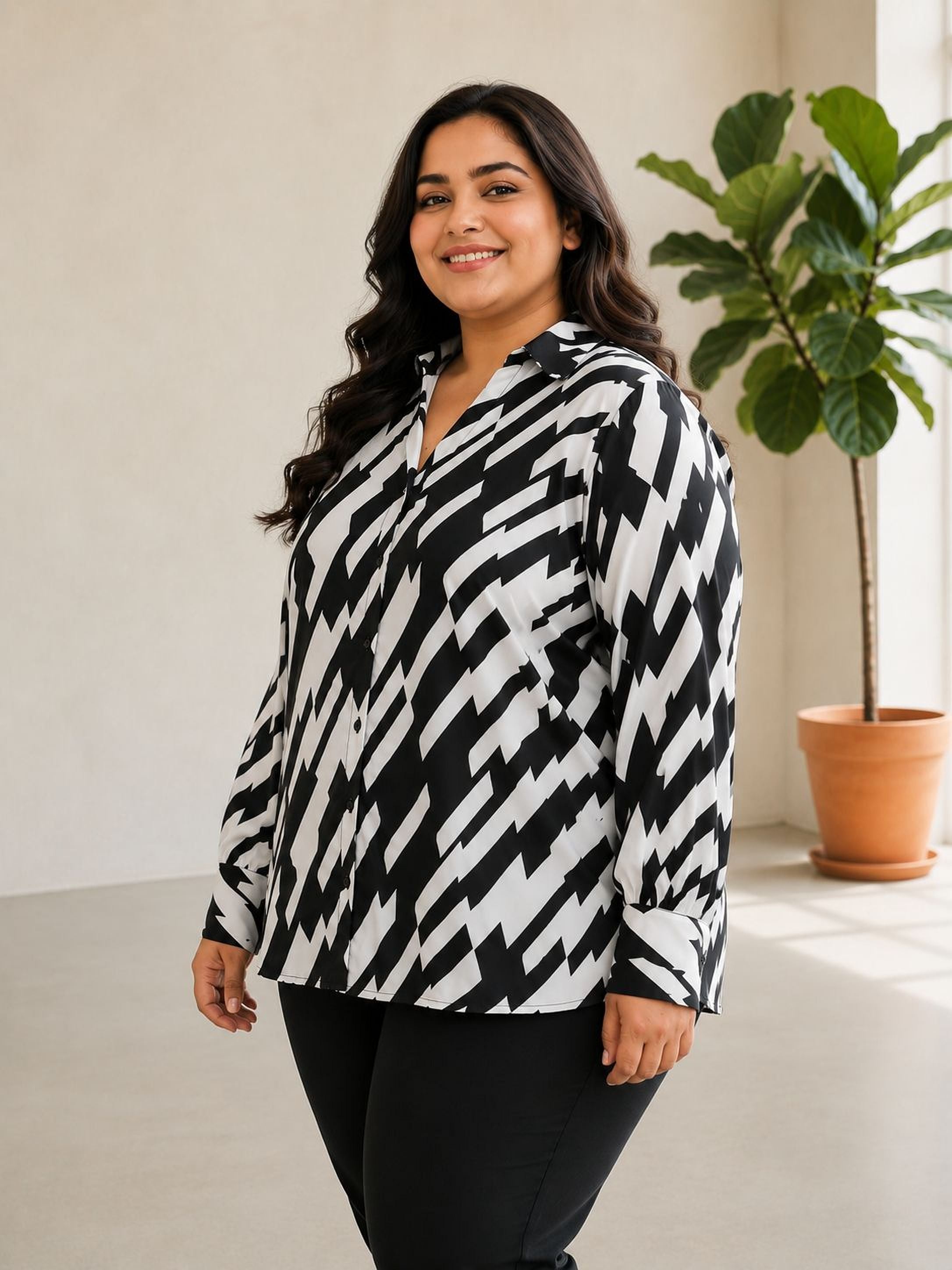 A woman with long dark hair is standing in a room, smiling at the camera. She is wearing a black and white patterned blouse and black pants. In the background, there is a potted plant.