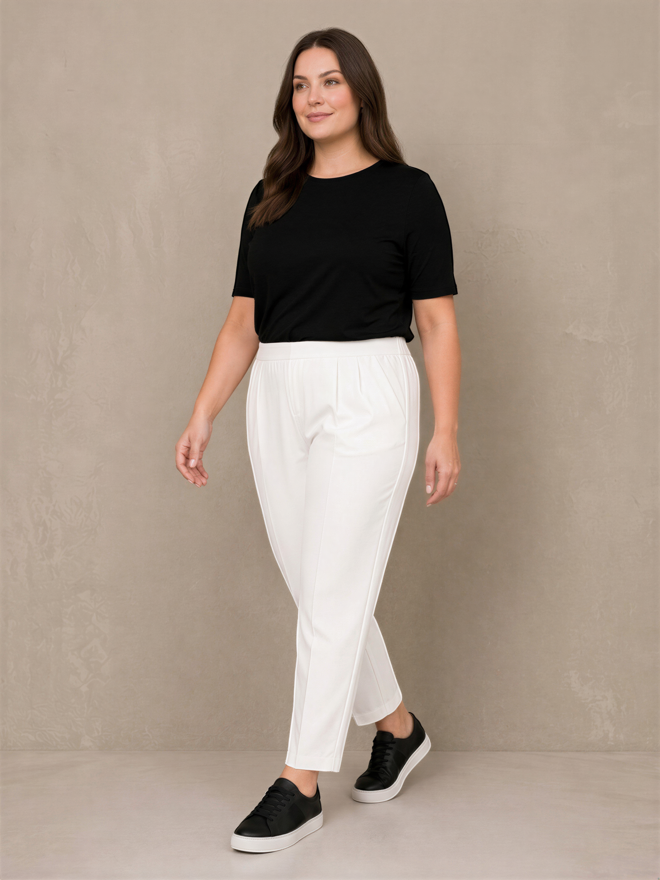 A woman wearing a black top and white pants stands against a beige wall, smiling at the camera.
