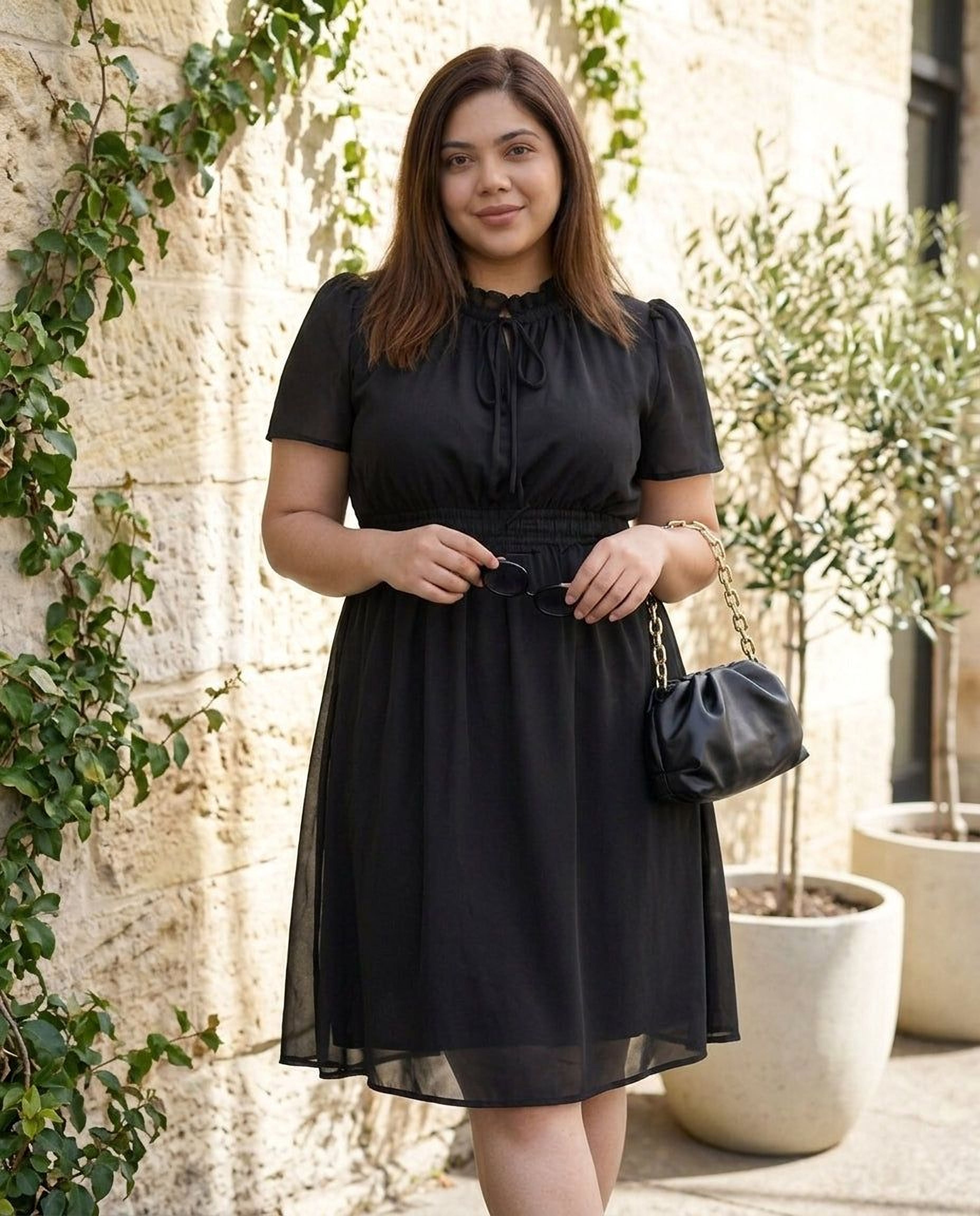 A woman wearing a black dress and high heels is smiling and looking down at her hand.