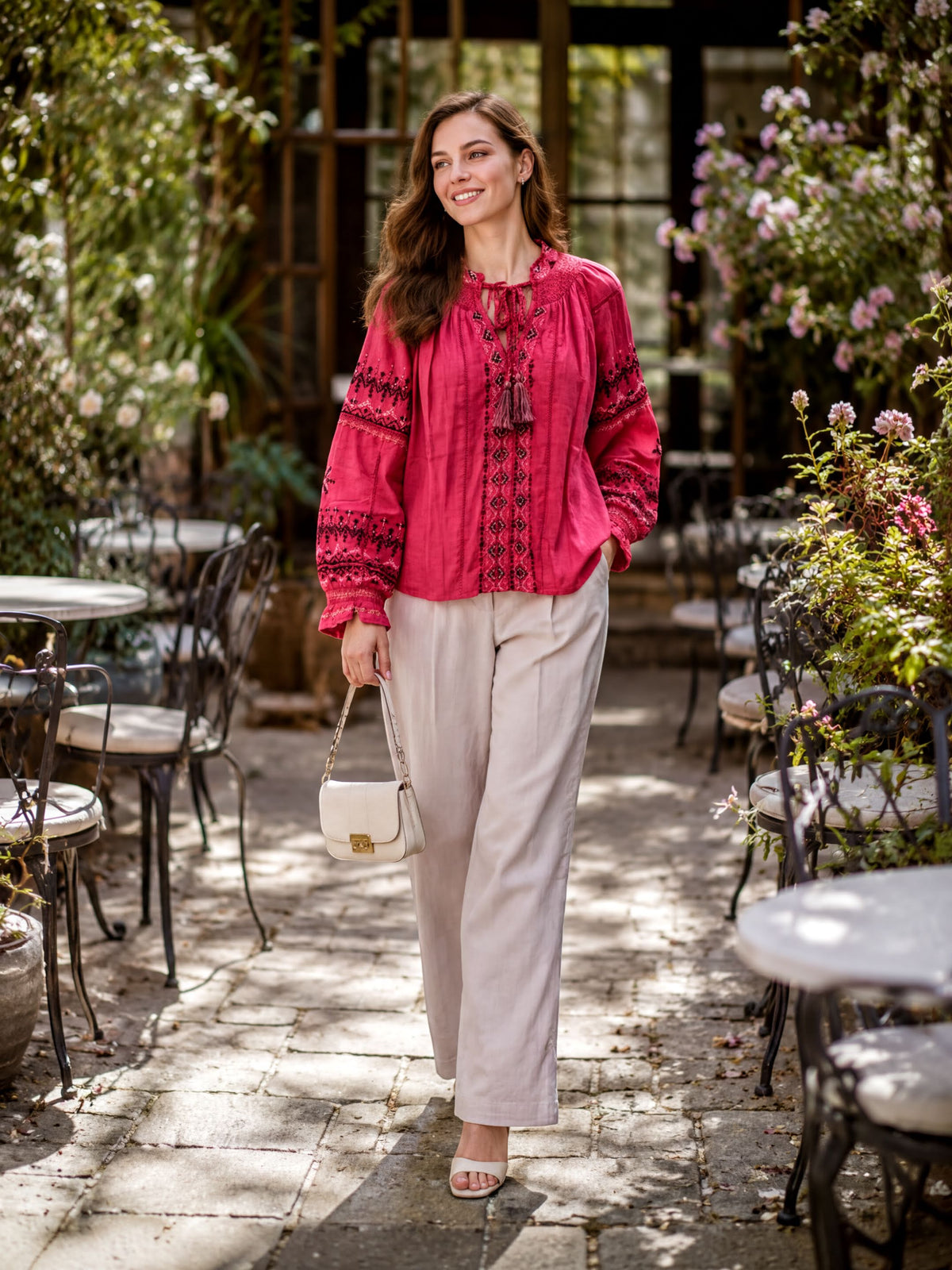 A woman in a red blouse and beige pants is walking down a path surrounded by tables and chairs, with flowers and greenery in the background.