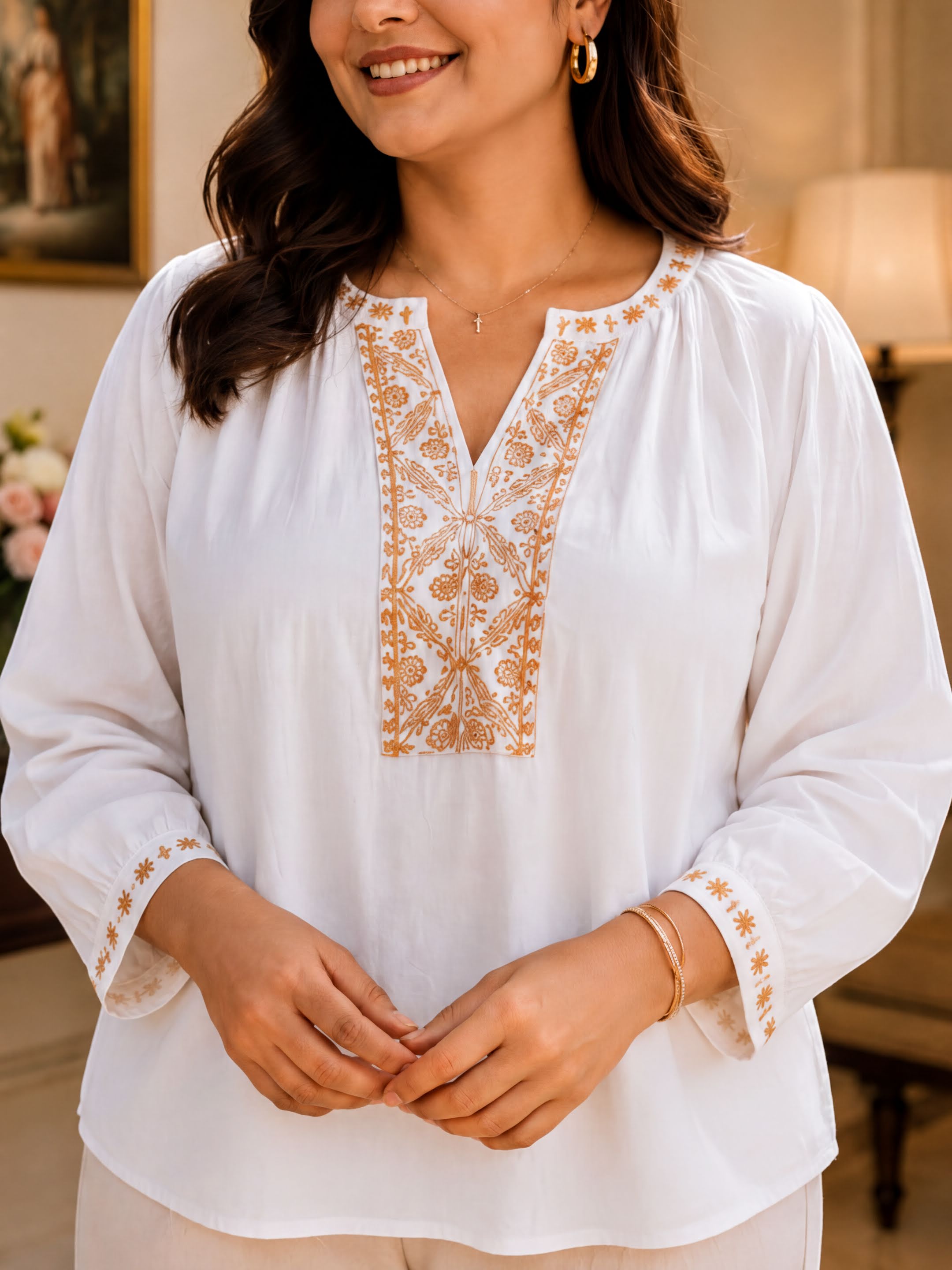 A woman wearing a white blouse with an embroidered pattern on the front, smiling and posing for the camera.
