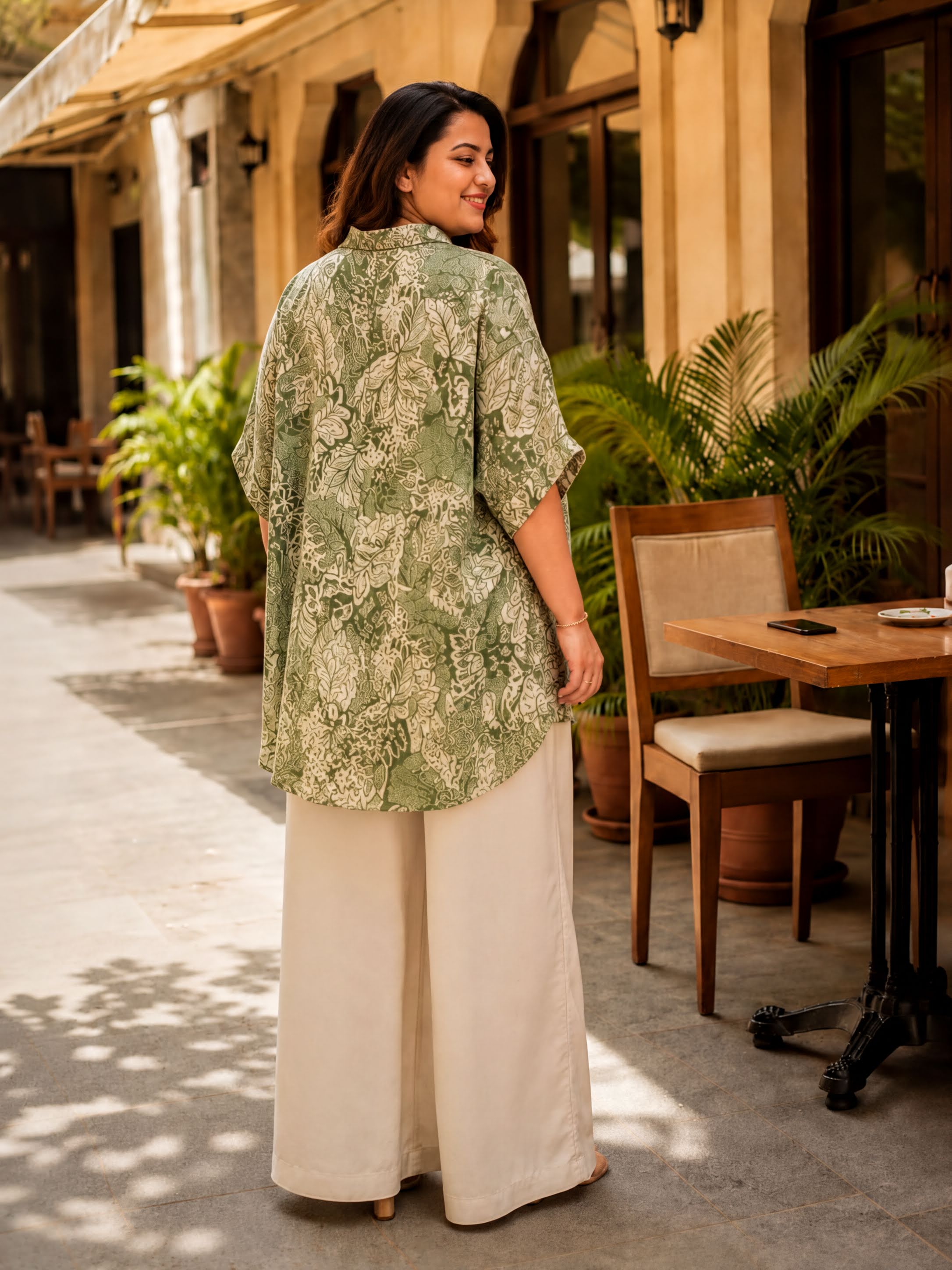 A woman wearing a green and white patterned blouse and white pants stands in front of a building with potted plants and a wooden table.