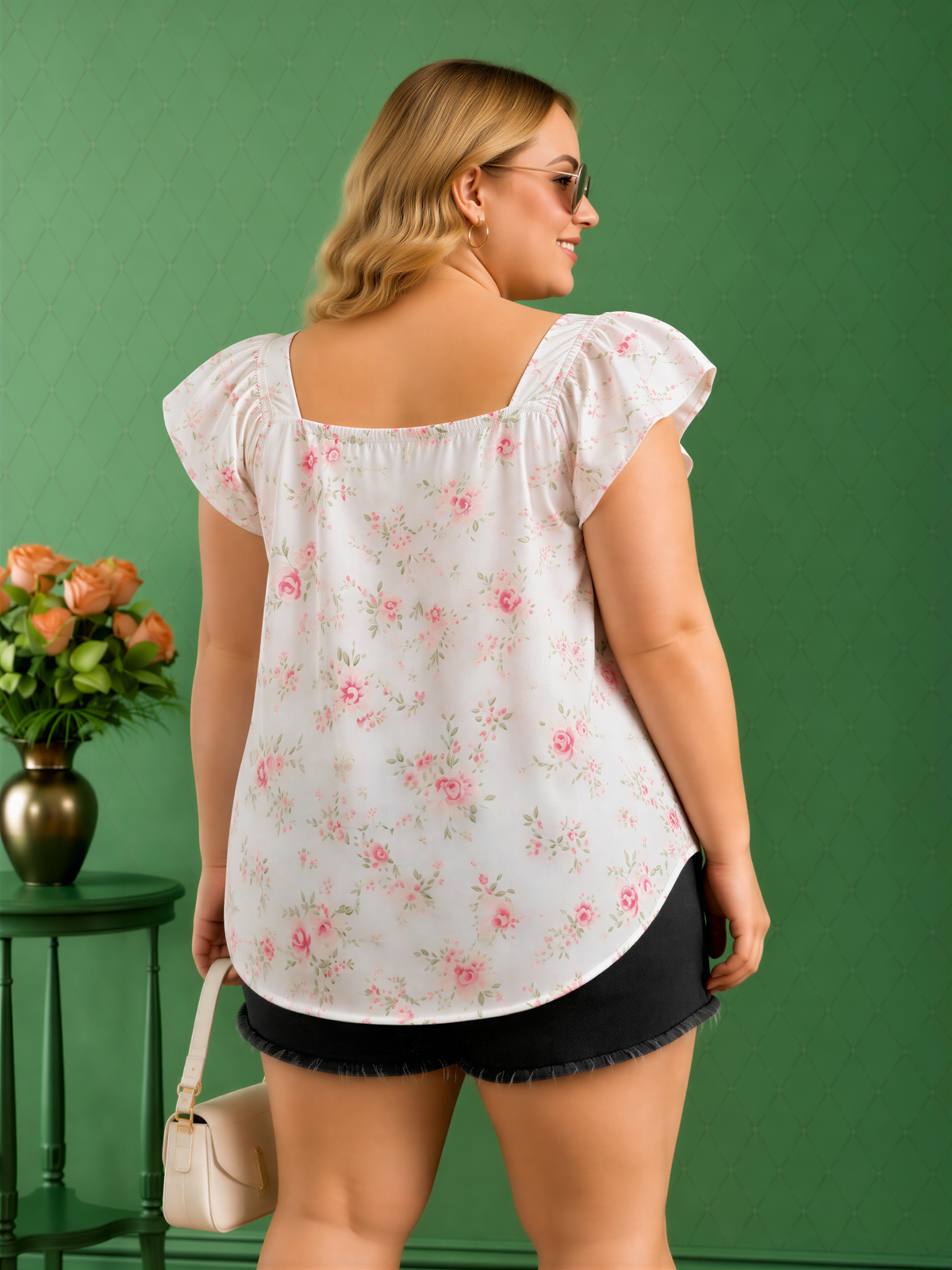 A woman wearing a white floral blouse and black shorts stands in front of a green wall, holding a small white purse.