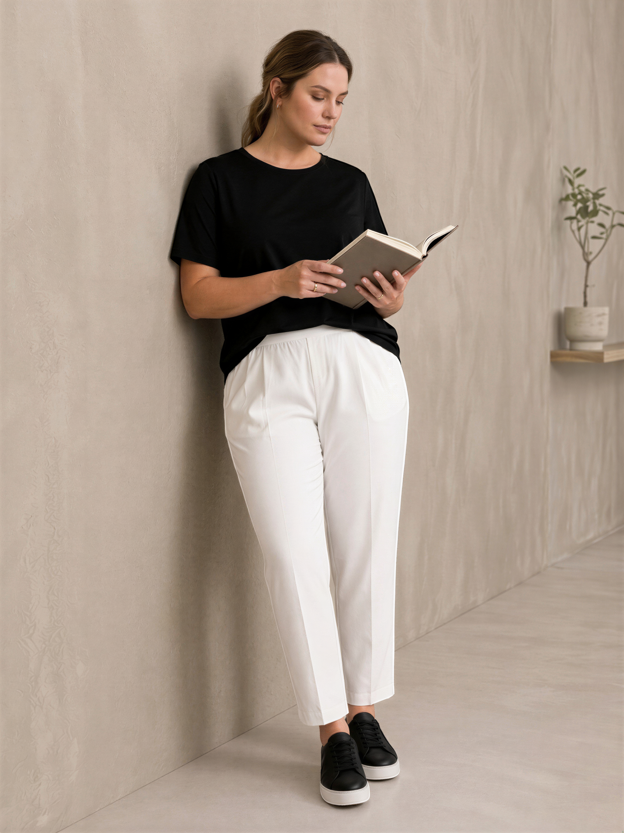 A woman in a black top and white pants is leaning against a wall and reading a book.