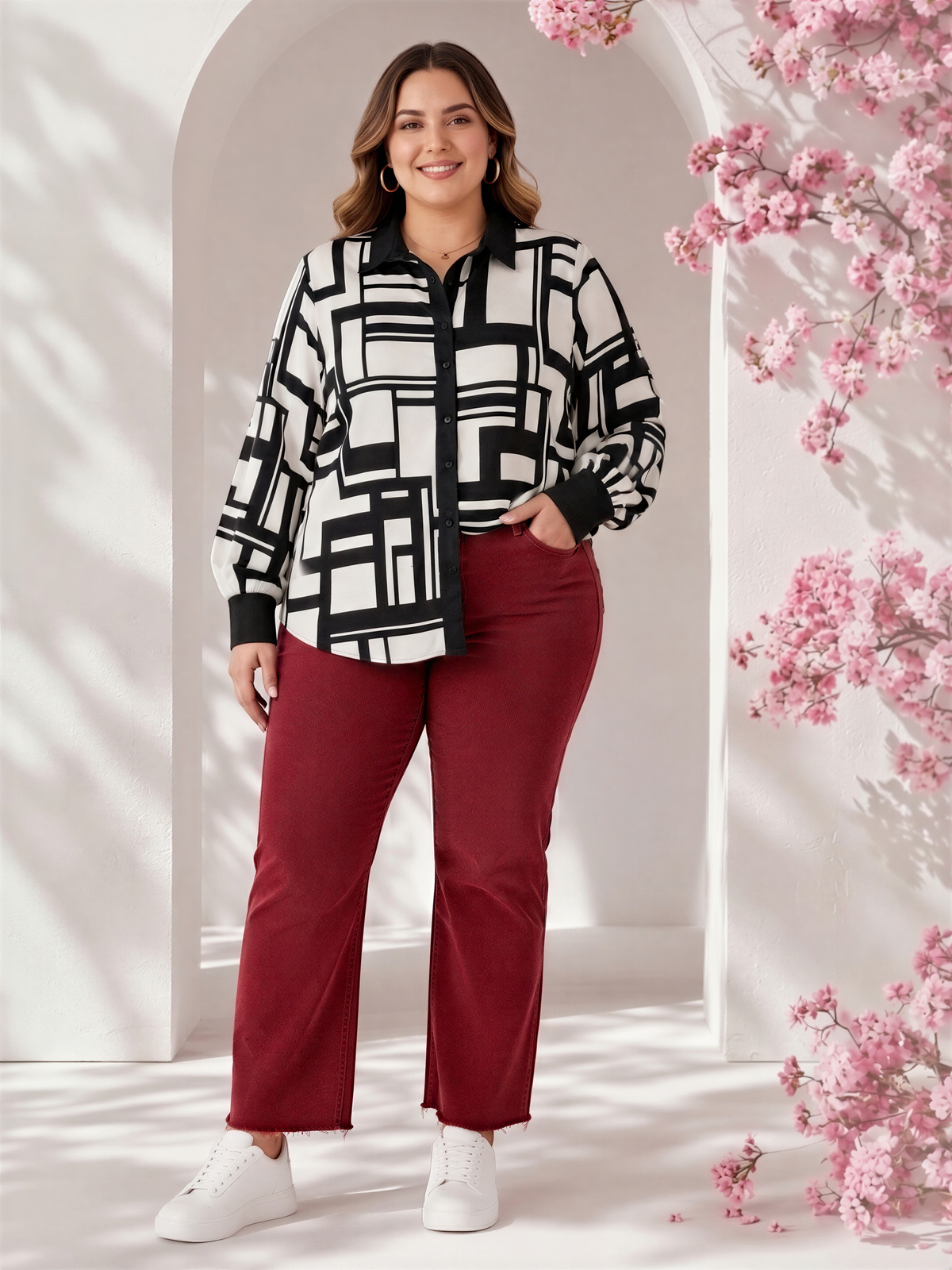 A woman wearing a black and white patterned blouse and red pants stands in front of a floral archway, smiling at the camera.
