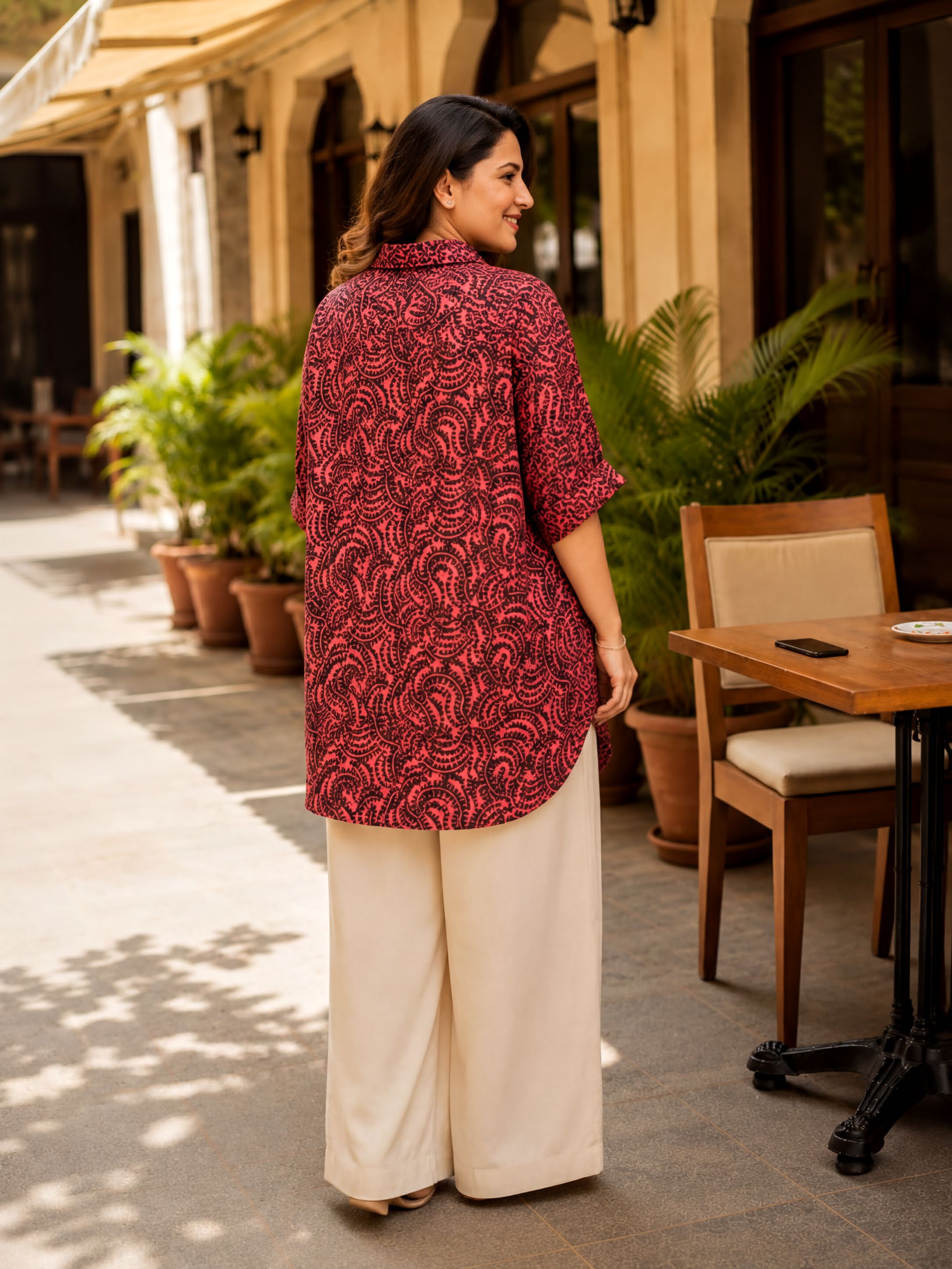 A woman wearing a red and black patterned blouse and beige wide leg pants stands in front of a building with potted plants and a wooden table.