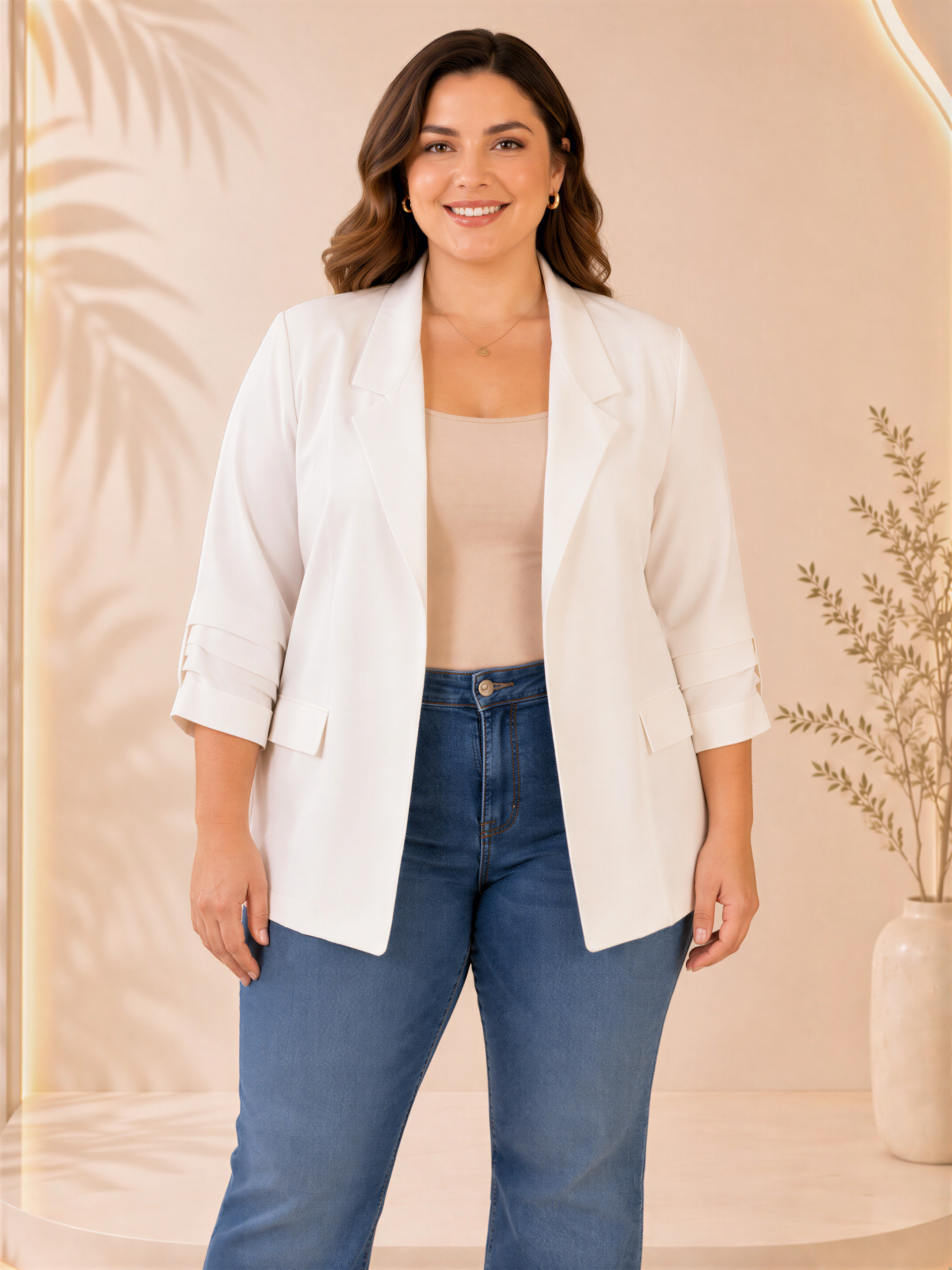 A woman wearing a white blazer, beige top, and blue jeans stands in front of a floral backdrop, smiling at the camera.