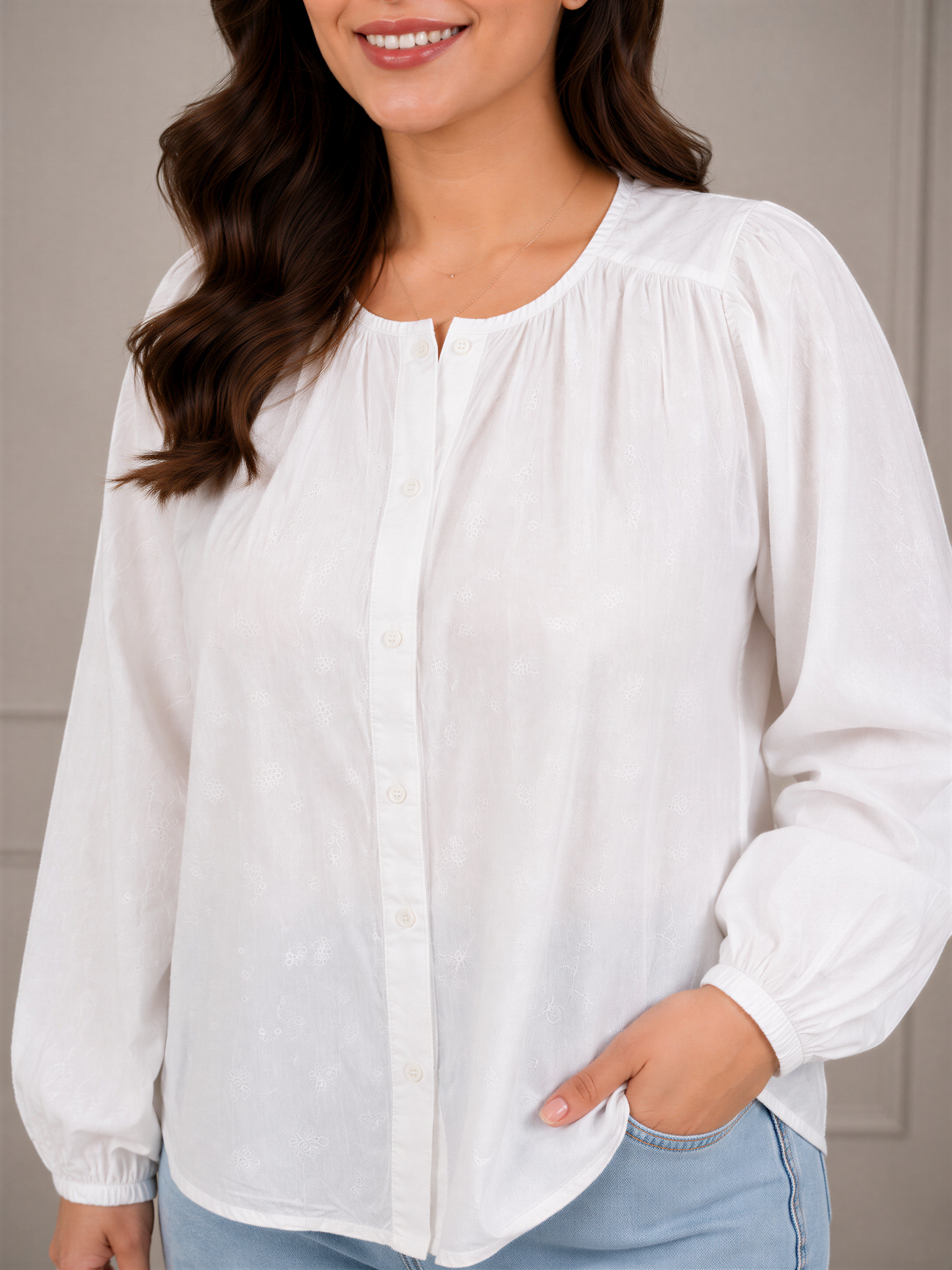 A woman with long brown hair wearing a white blouse and blue jeans is smiling and looking to the side.