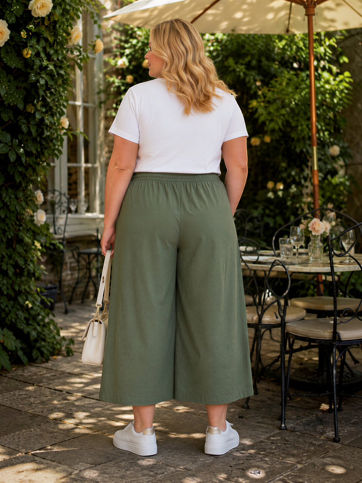 A woman wearing a white t-shirt, olive green wide-leg pants, and white sneakers stands in front of a table with a vase of flowers and a white umbrella.
