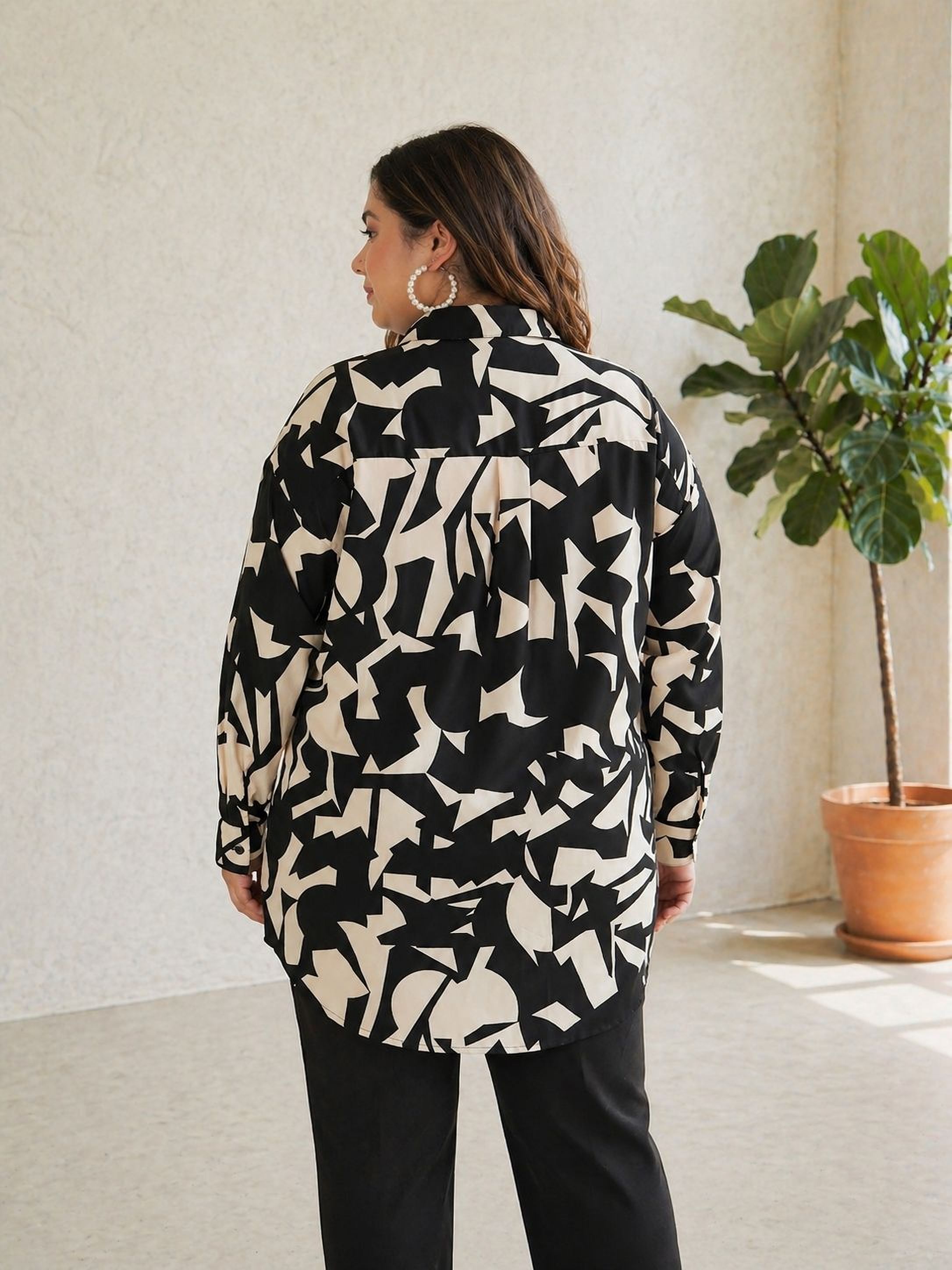 A woman wearing a black and white patterned shirt stands in a room with a potted plant in the background.