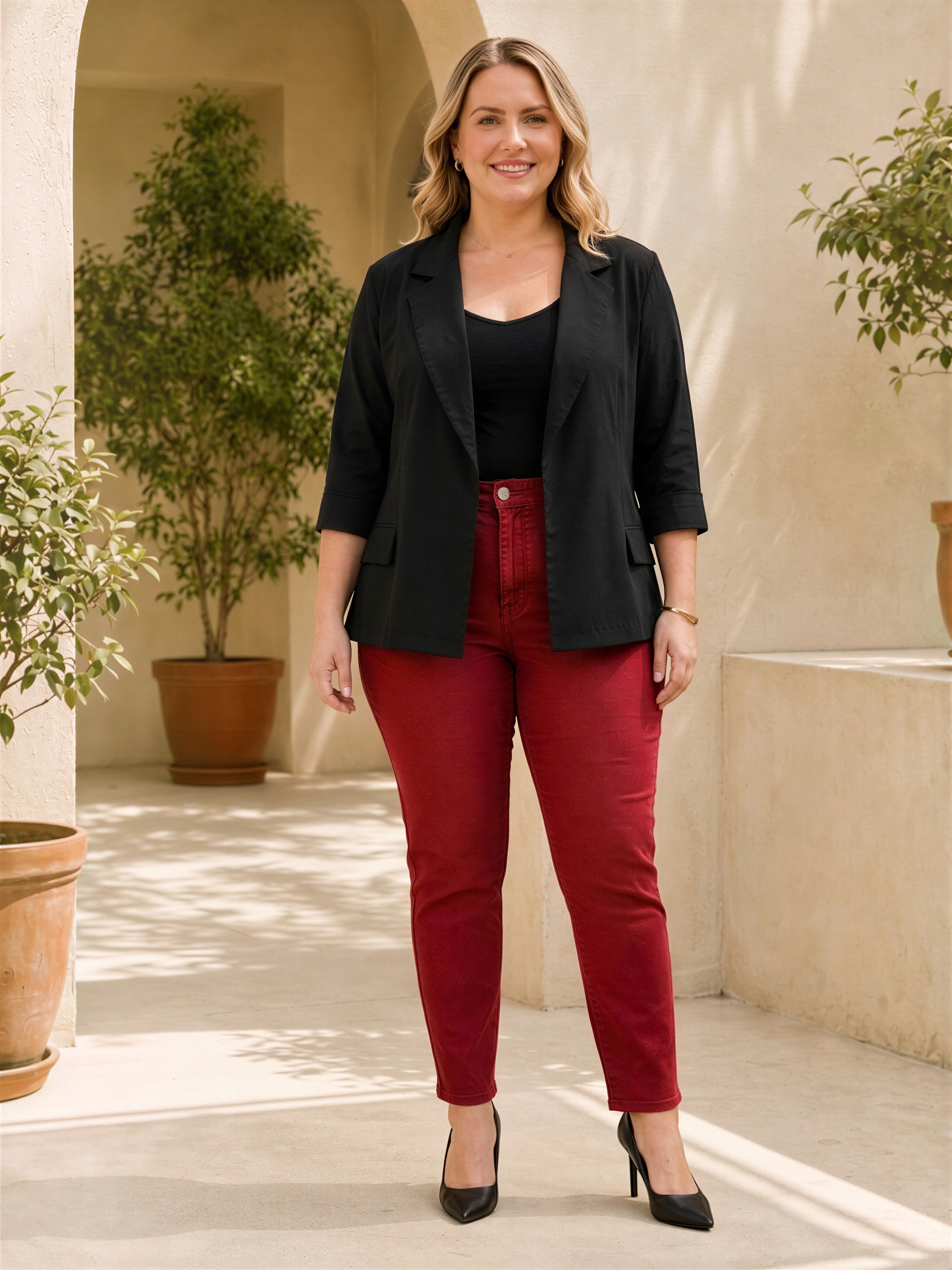 A woman wearing a black blazer, red pants, and black heels stands in a hallway with potted plants in the background.