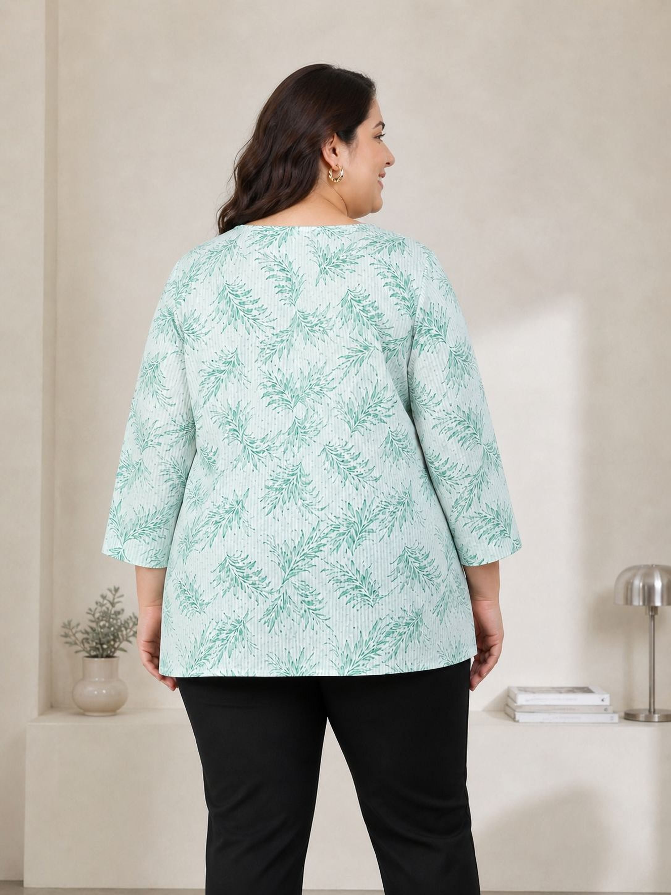 A woman wearing a light green blouse with a leaf pattern stands in front of a white wall, smiling at the camera.