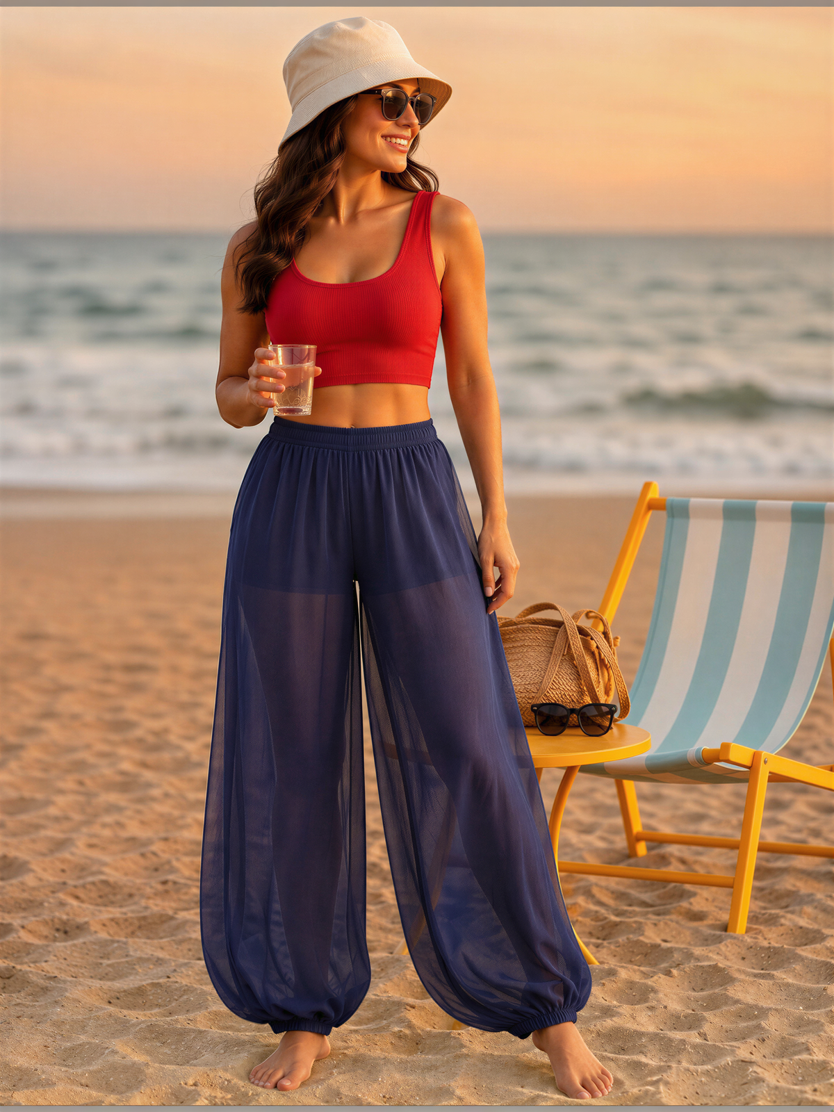 A woman in a red crop top and blue pants stands on a beach, holding a drink and wearing a white hat.
