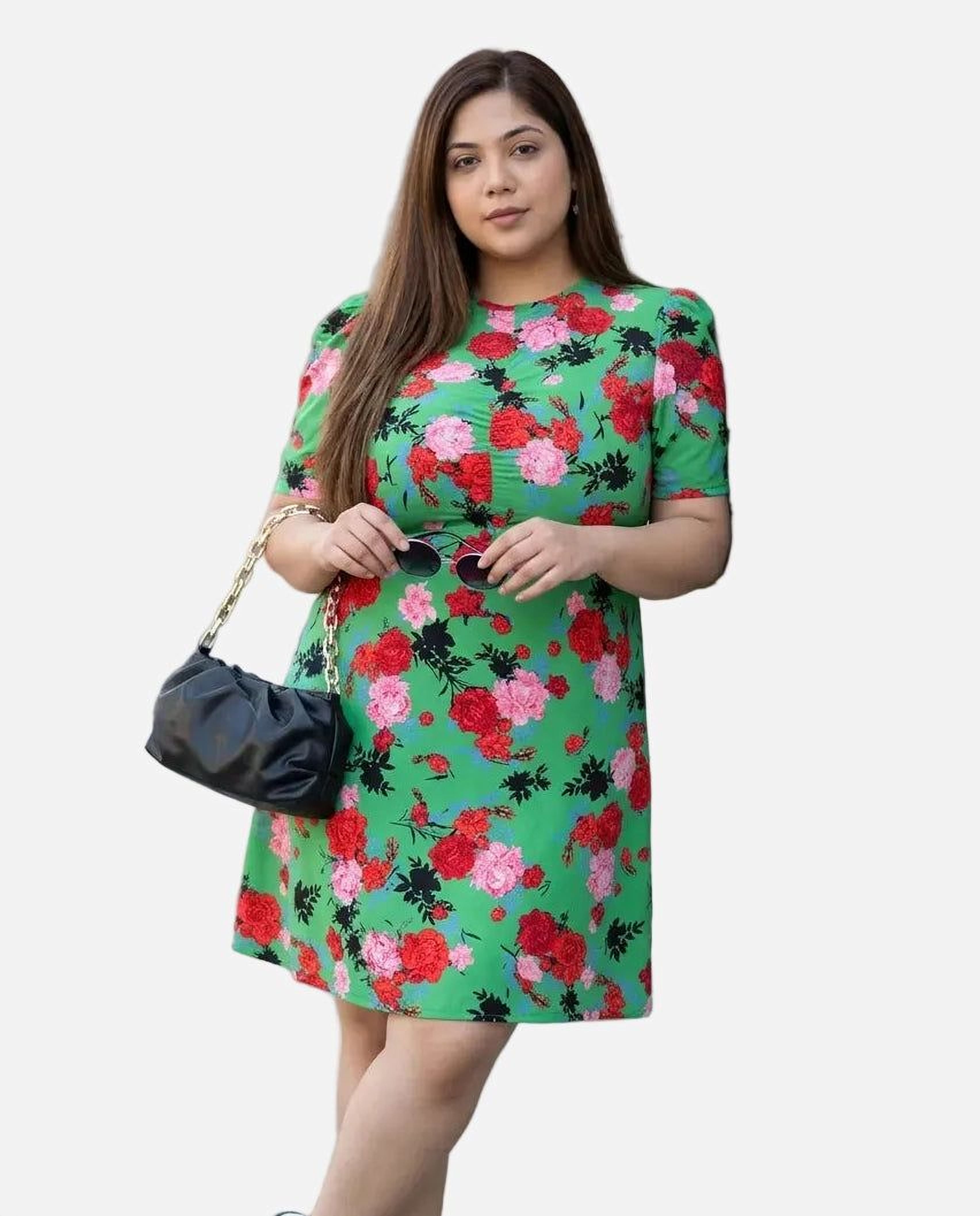 A woman wearing a green floral print dress and sunglasses, holding a black purse, and smiling at the camera.
