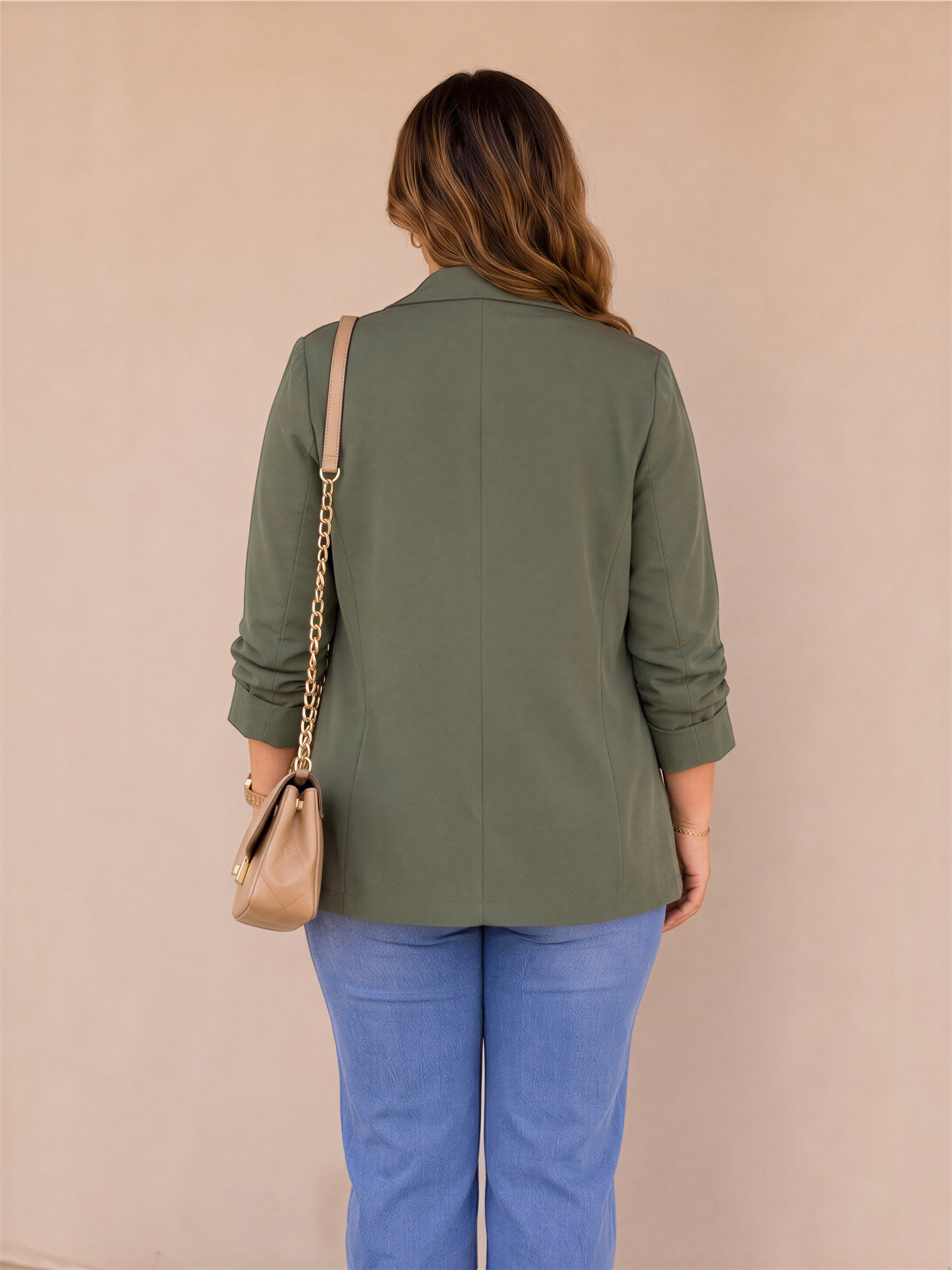 A woman wearing a green blazer and blue jeans stands facing away from the camera, with a beige purse hanging from her shoulder.