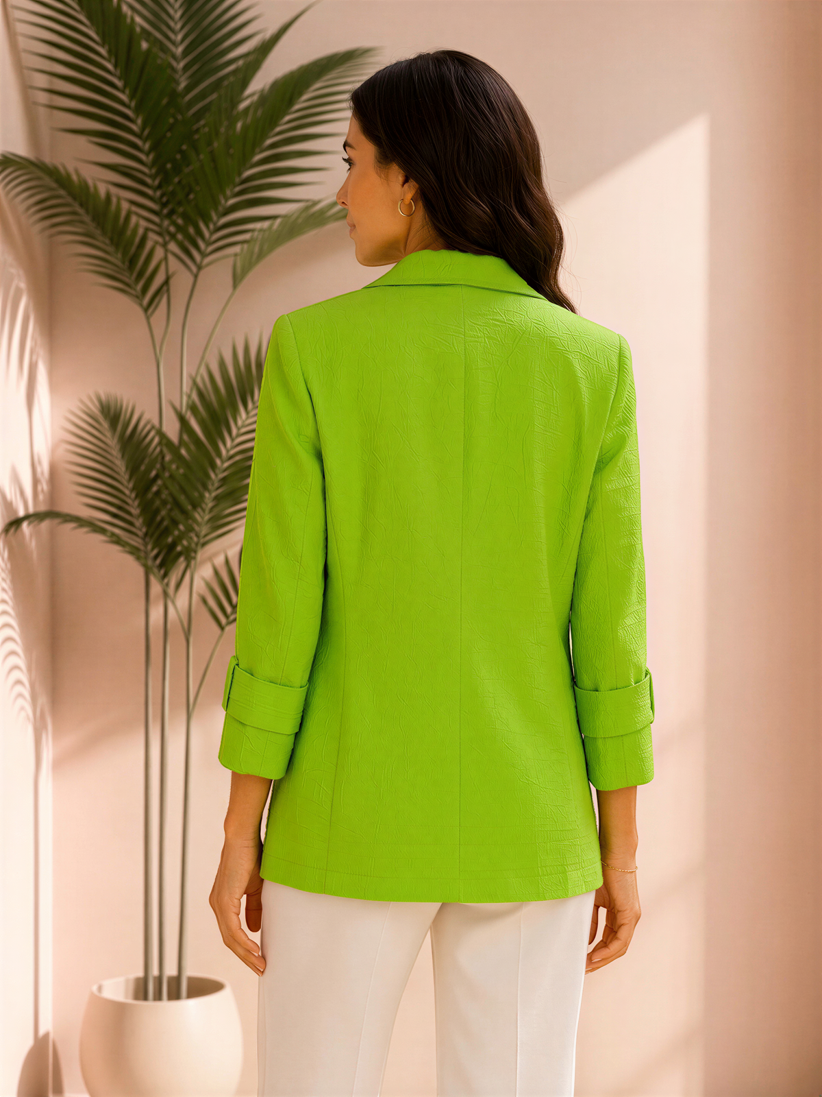 A woman wearing a bright green blazer stands in front of a plant, facing away from the camera.