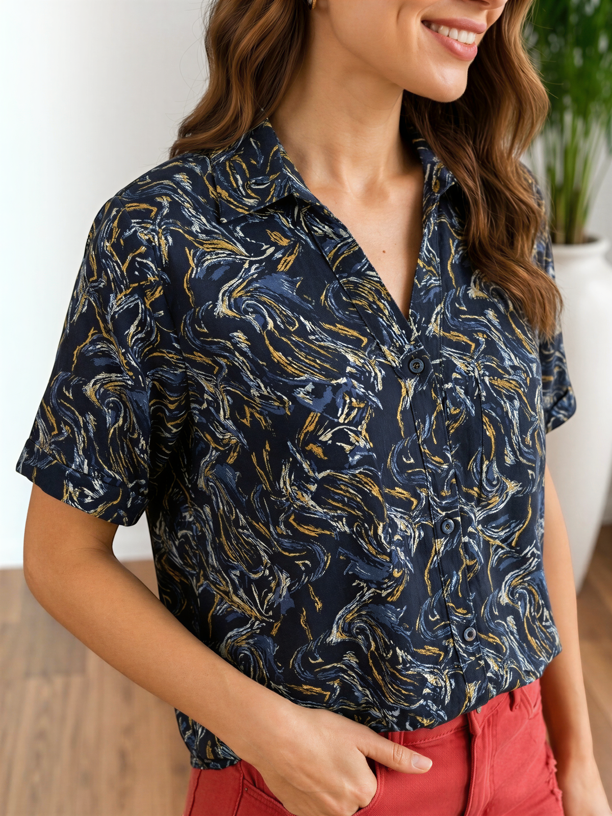 A woman wearing a blue and gold patterned shirt and red pants is smiling and looking to the side.