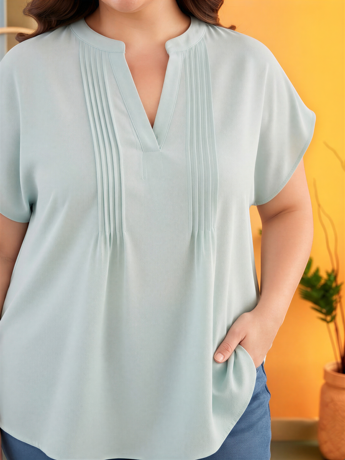 A woman wearing a light blue blouse with a v-neck and short sleeves stands in front of a yellow wall with a potted plant.
